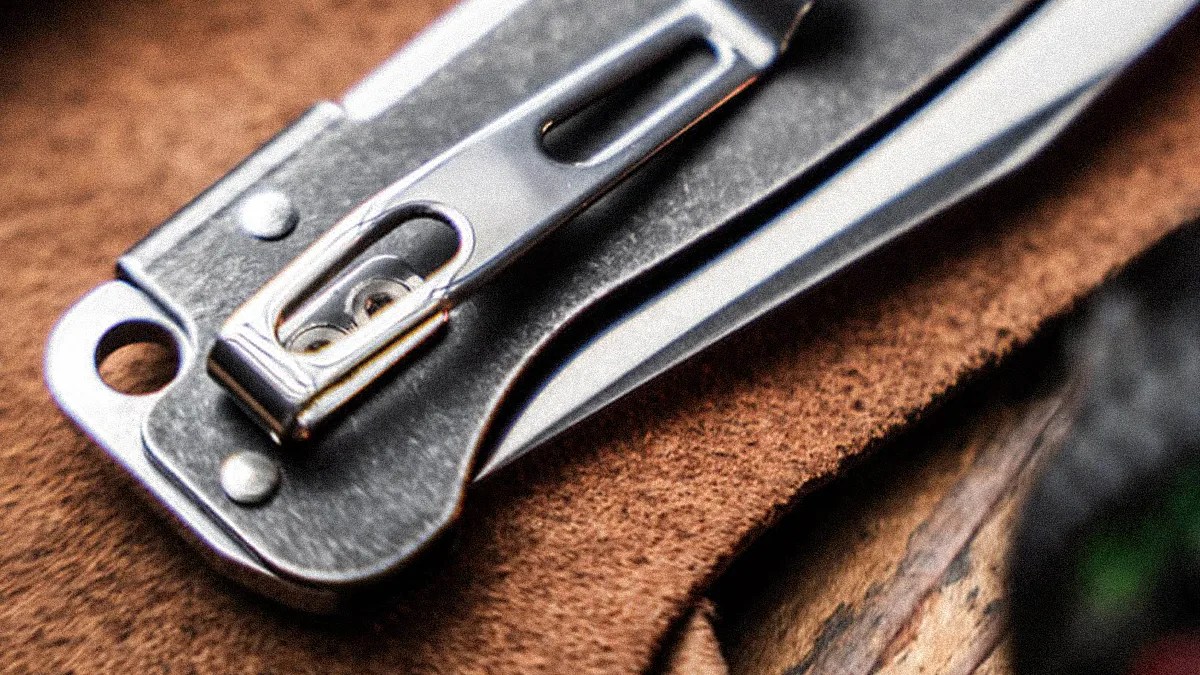 Close-up of a silver metal folding knife with a clip resting on brown suede material.