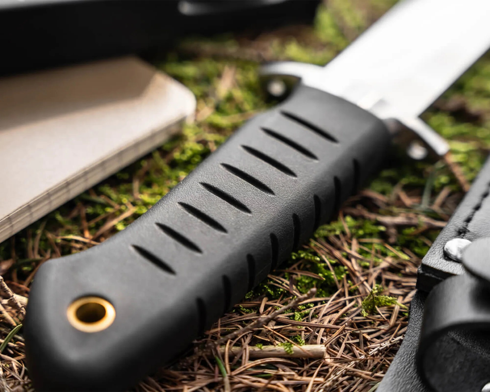 Black textured knife handle with grooves and a brass lanyard hole on forest floor.