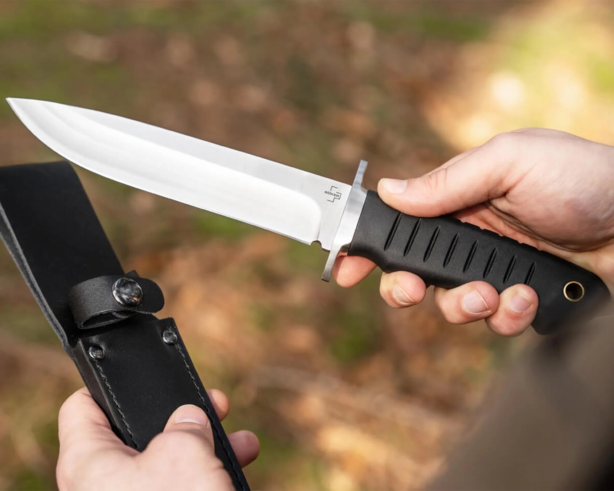 Hand holding a fixed-blade knife with a black textured handle and a silver blade next to a black leather sheath.