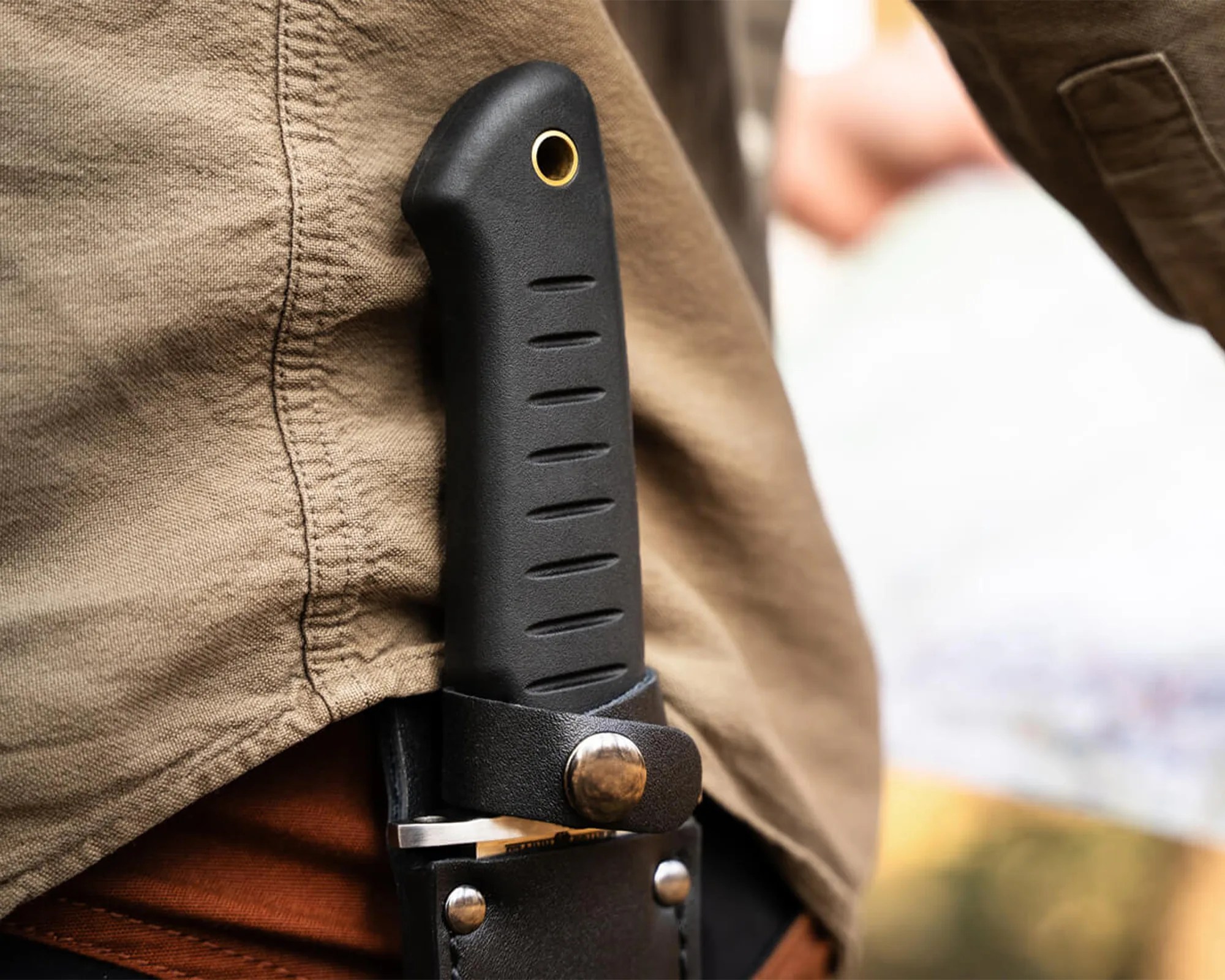 Black textured knife handle with a brass lanyard hole secured in a black leather sheath on a person's belt.