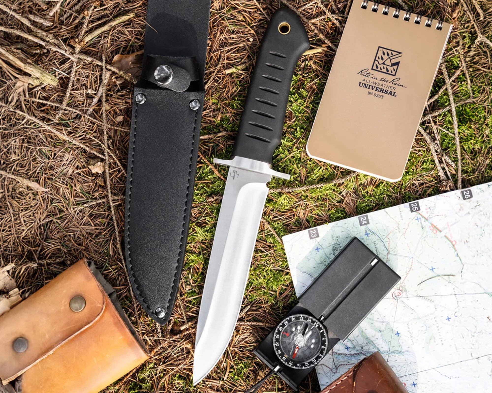 Hunting knife with black handle and sheath, compass, map, and tan all-weather notebook on forest floor.
