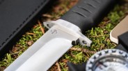 Close-up of a Böker fixed-blade knife with a black handle resting on green moss and pine needles.