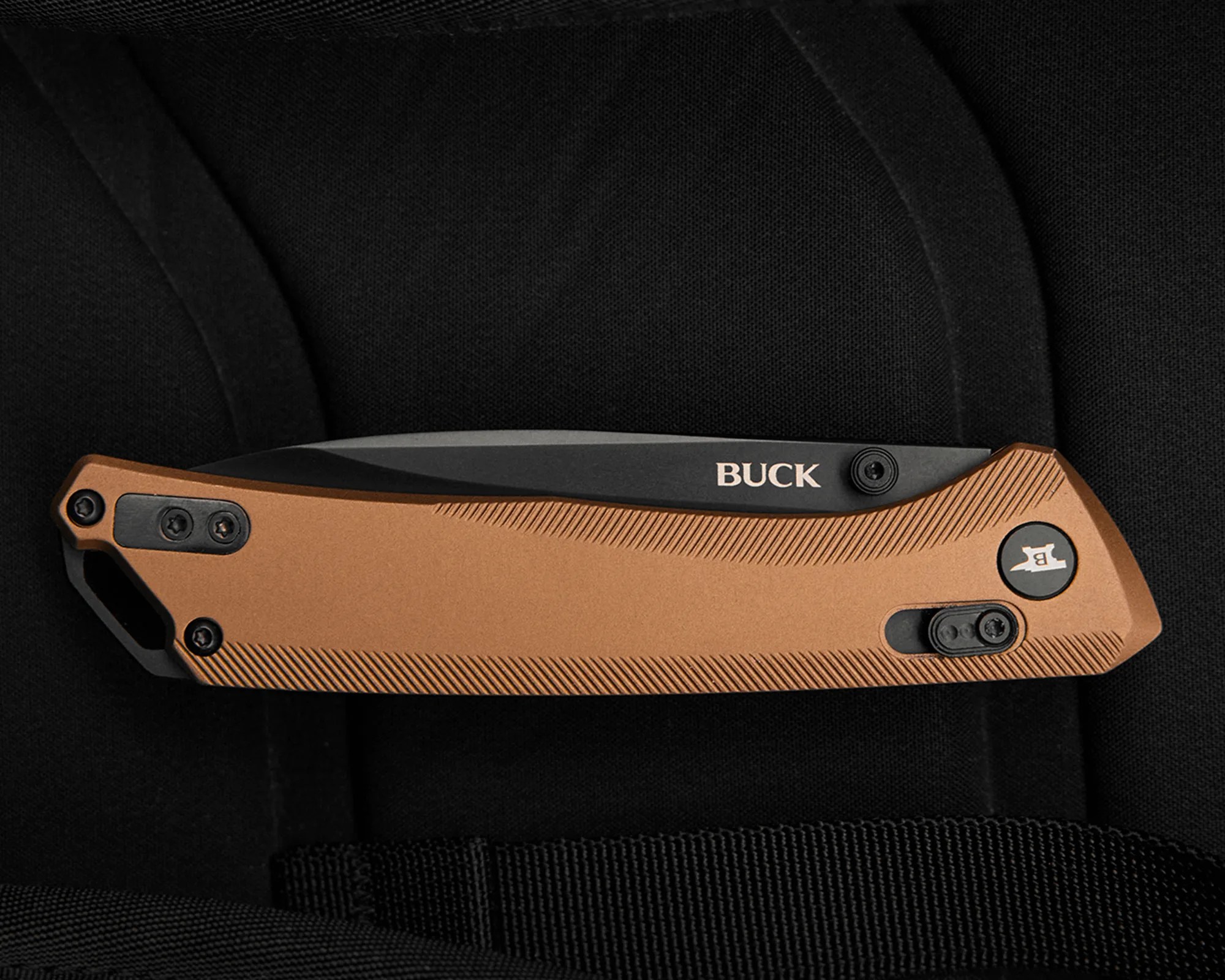 Closed folding knife with a copper-colored textured handle and black blade marked "BUCK" on a black fabric background.