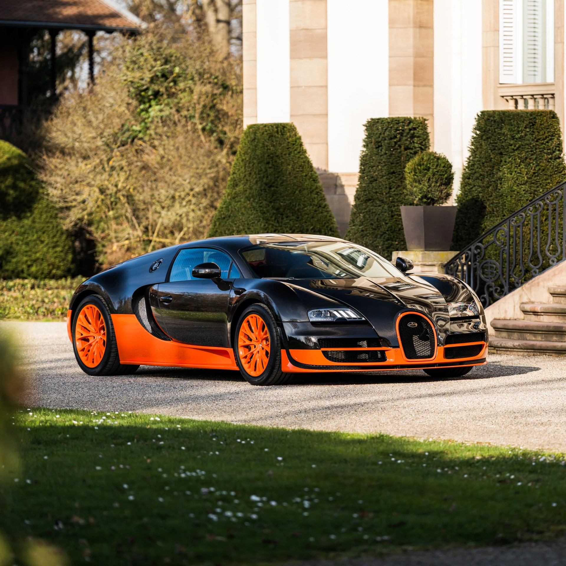 Black and orange Bugatti Veyron sports car parked on a gravel driveway in front of manicured bushes and a stone building.