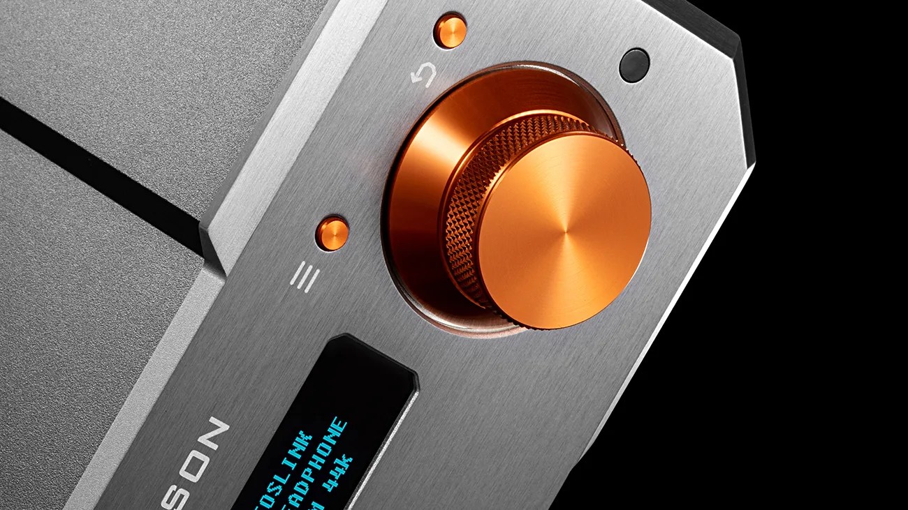 Close-up of a brushed metal audio device with an orange volume knob and a blue digital display.