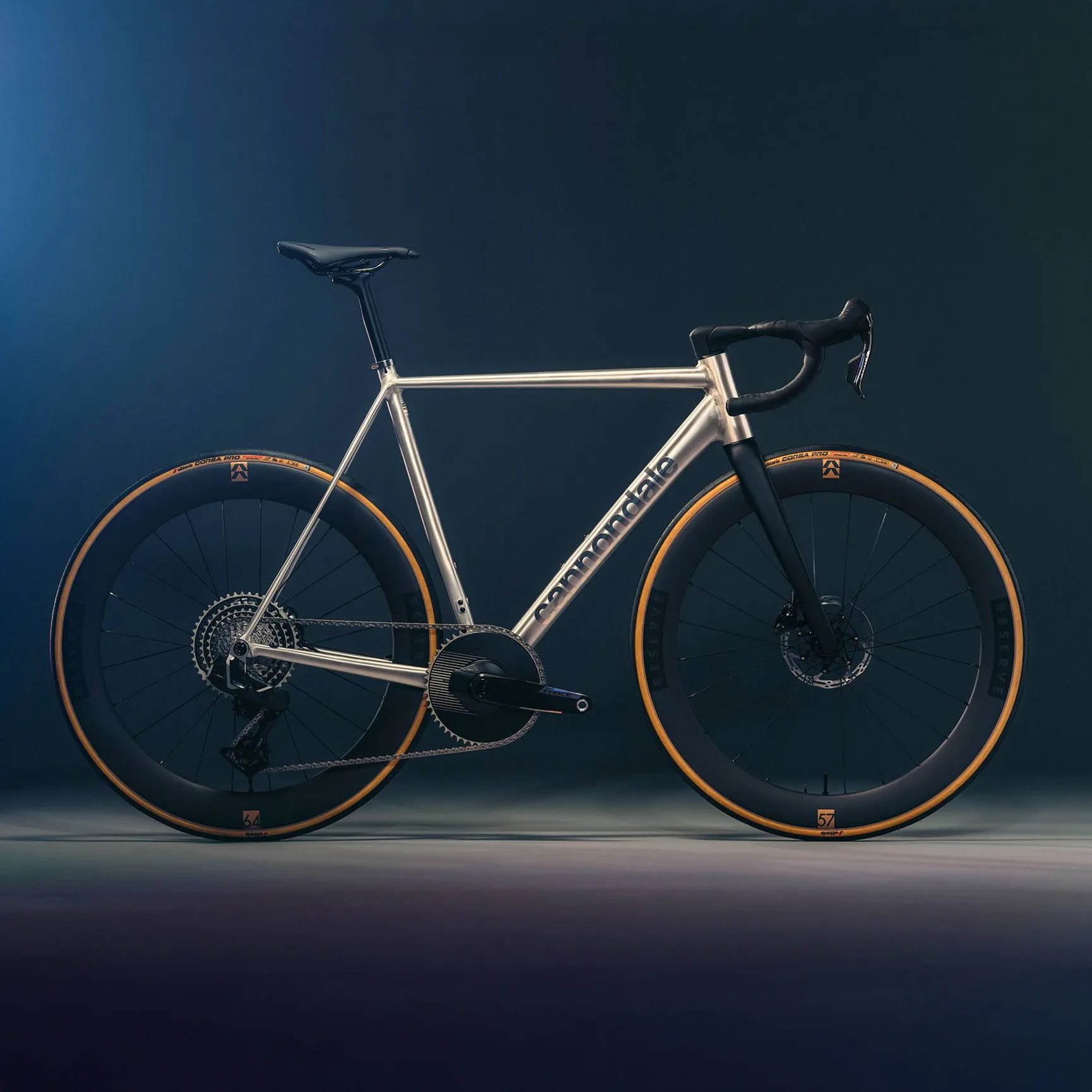 Silver Cannondale road bike with black deep-section wheels and tan sidewall tires on a dark background.