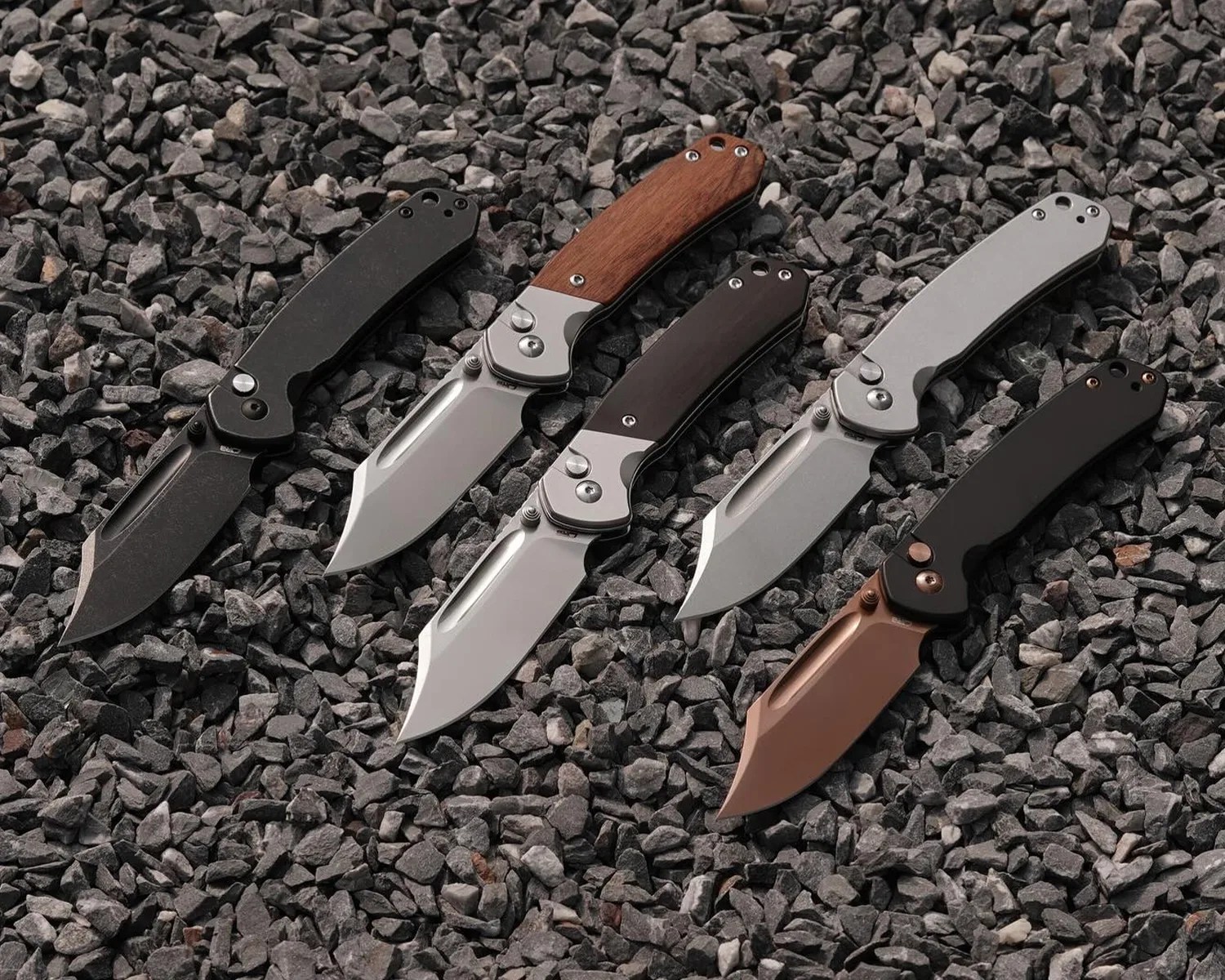 Five folding knives with different handle colors and blade finishes laid out on dark gray gravel.