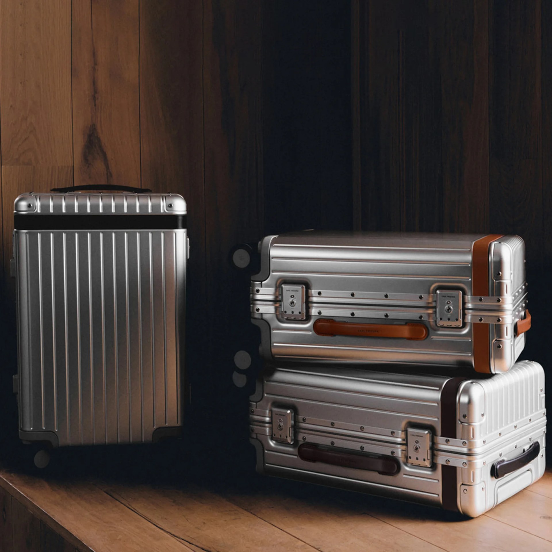 Three silver hard-shell suitcases with vertical ridges, two stacked horizontally with brown and black leather handles, and one standing upright.
