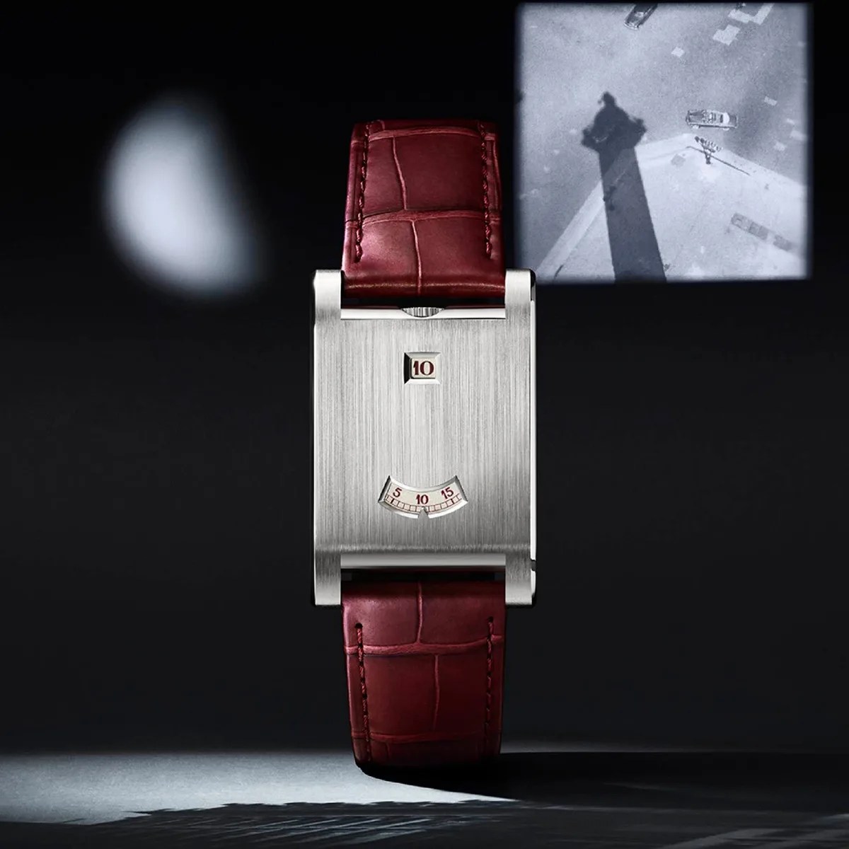 Rectangular silver watch with a brushed metal face and burgundy leather strap, showing digital hour and minute windows.