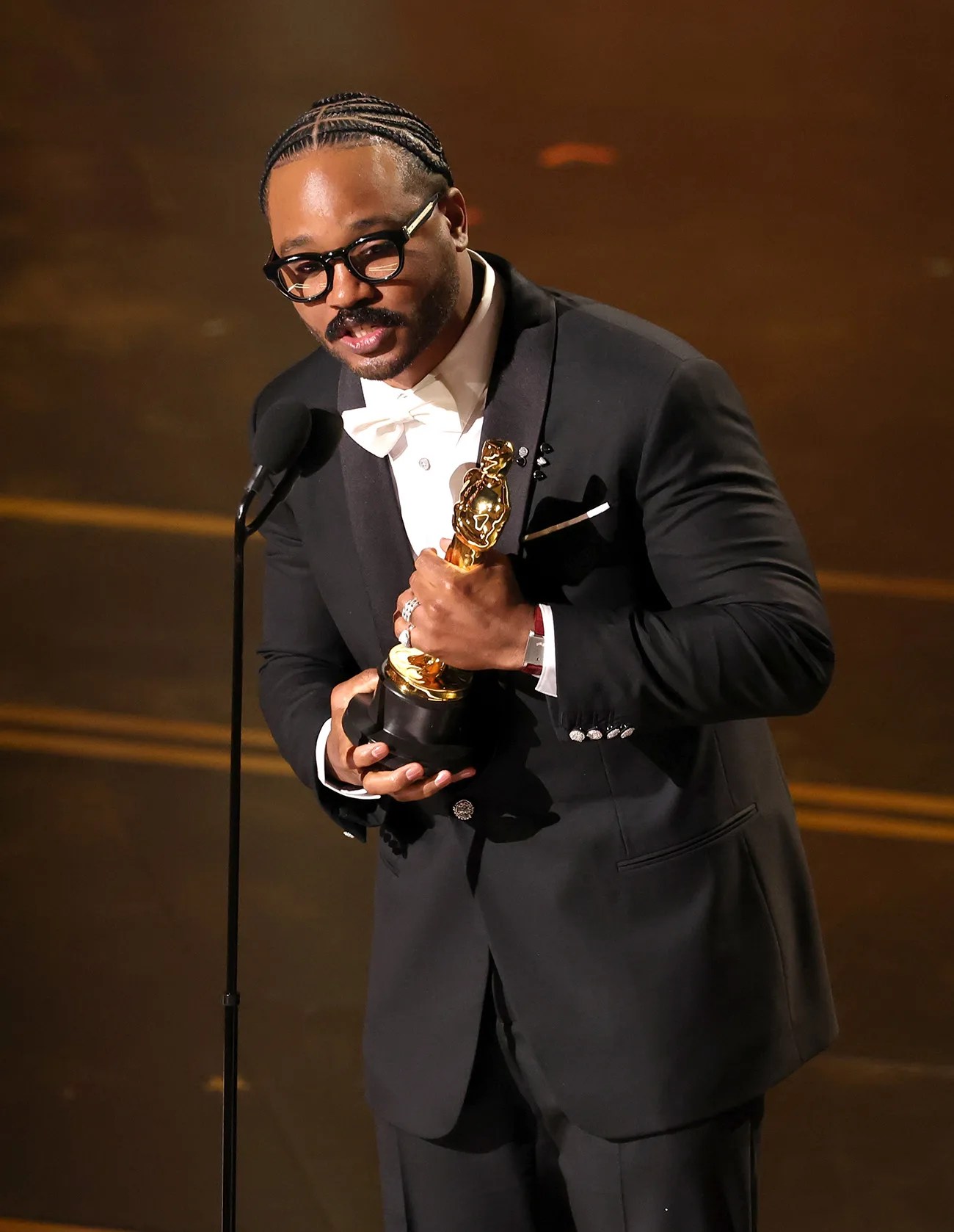 Man in black tuxedo with white bow tie holding an Oscar statuette at a microphone on stage.