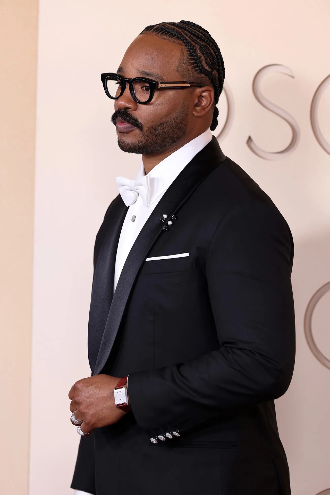 Man wearing a black tuxedo with satin lapels, white bow tie, black glasses, and a red leather strap watch.
