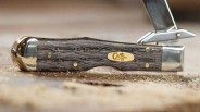 Folding pocket knife with dark wood handle, brass rivets, and partially open metal blade on a sandy surface.