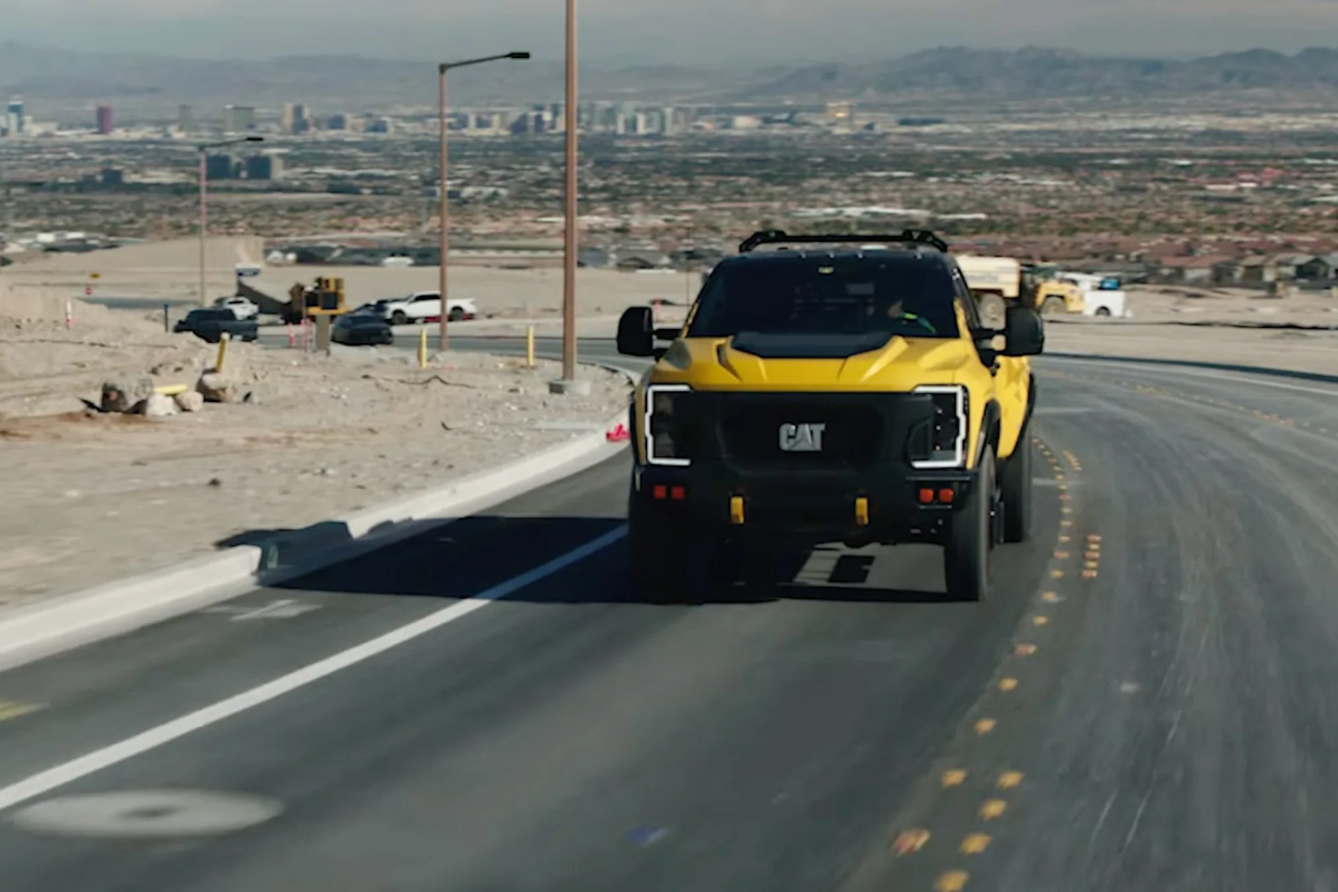 Yellow and black CAT electric pickup truck driving on a paved road with a desert cityscape in the background.