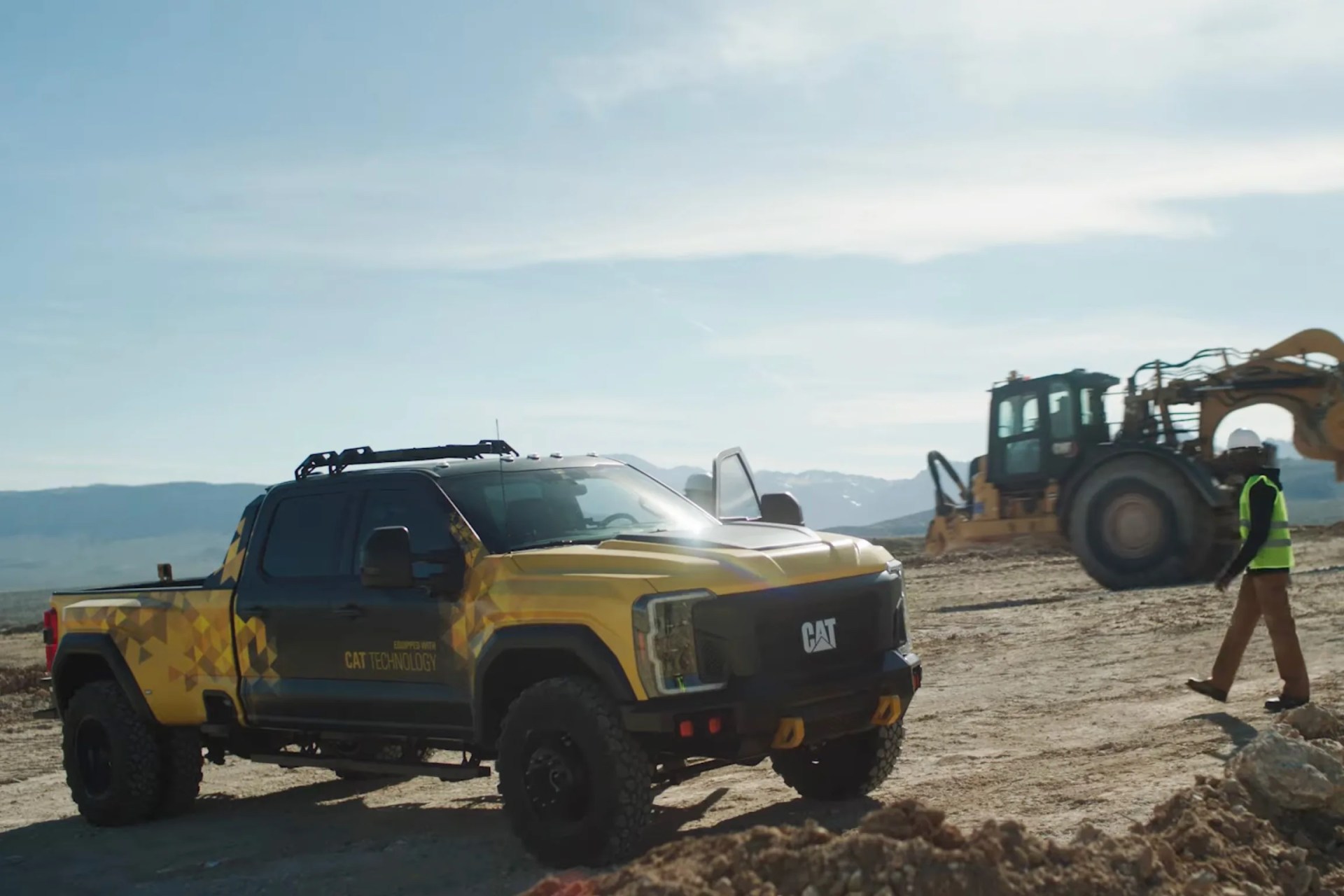 Yellow and black CAT pickup truck with off-road tires parked on a dirt construction site with heavy machinery in the background.