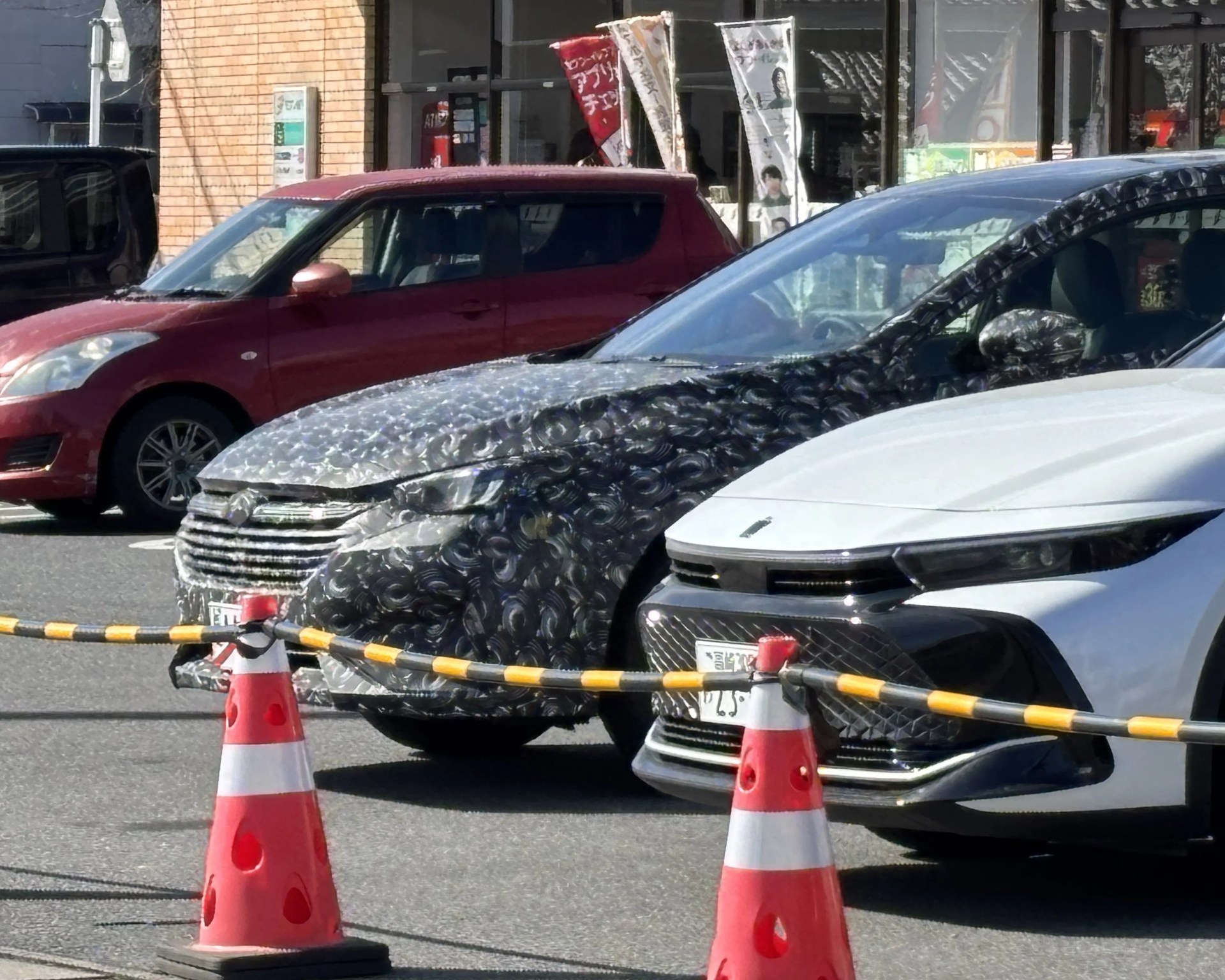 Two cars covered in black and silver camouflage wrap parked next to a white car behind orange traffic cones and a yellow-black striped barrier.