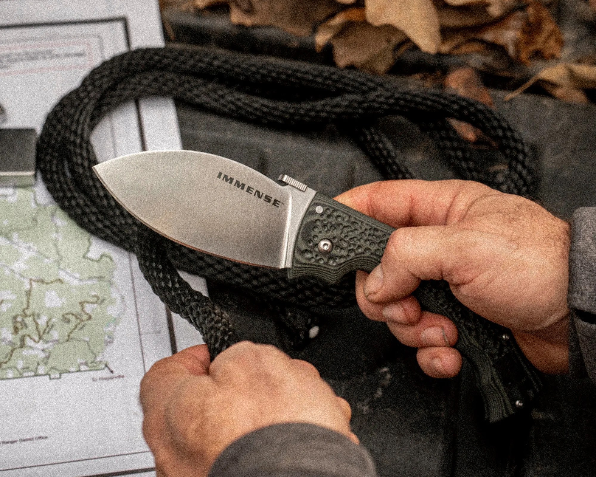 Person holding a fixed-blade knife with "IMMENSE" on the blade and a textured dark green handle, cutting black rope.