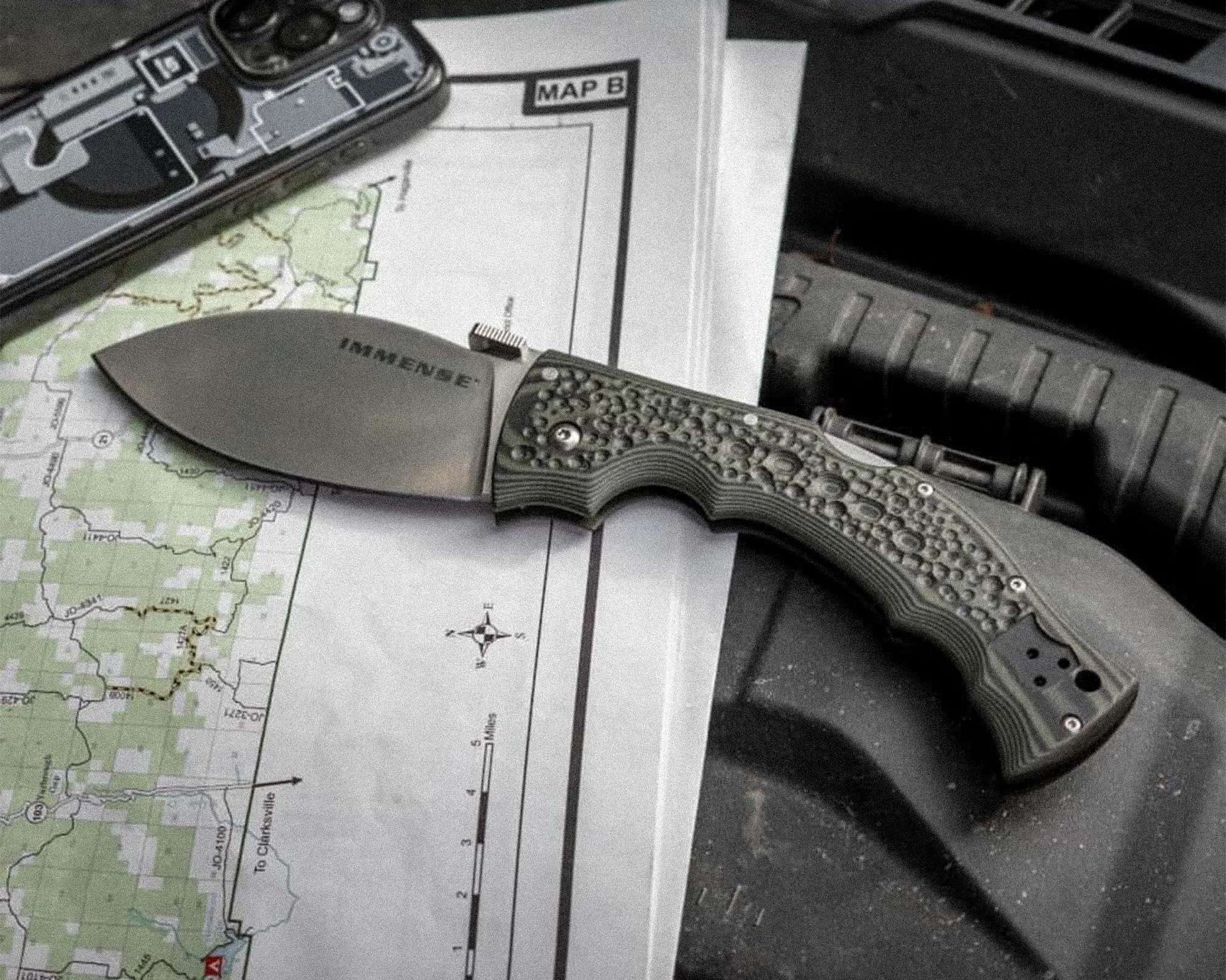 Folding knife with textured green handle and "IMMENSE" engraved on the blade, resting on a map and near a smartphone.