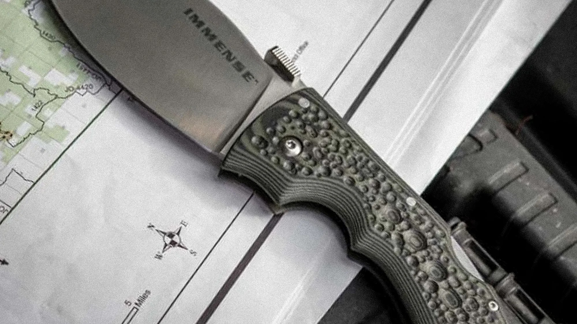 Folding knife with textured green handle and partially visible blade marked "EMERGENCE" on a map.