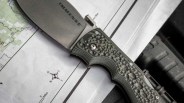 Folding knife with textured green handle and partially visible blade marked "EMERGENCE" on a map.