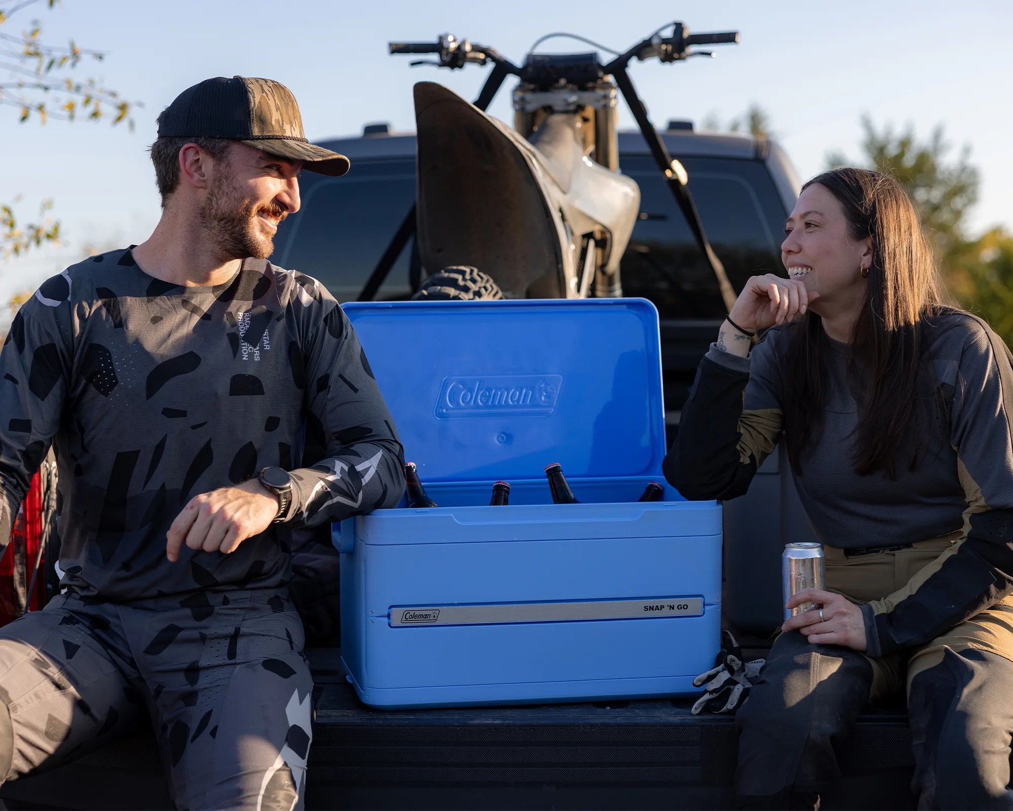 Blue Coleman Snap 'N Go cooler with bottles inside on a truck bed between two people sitting and smiling.