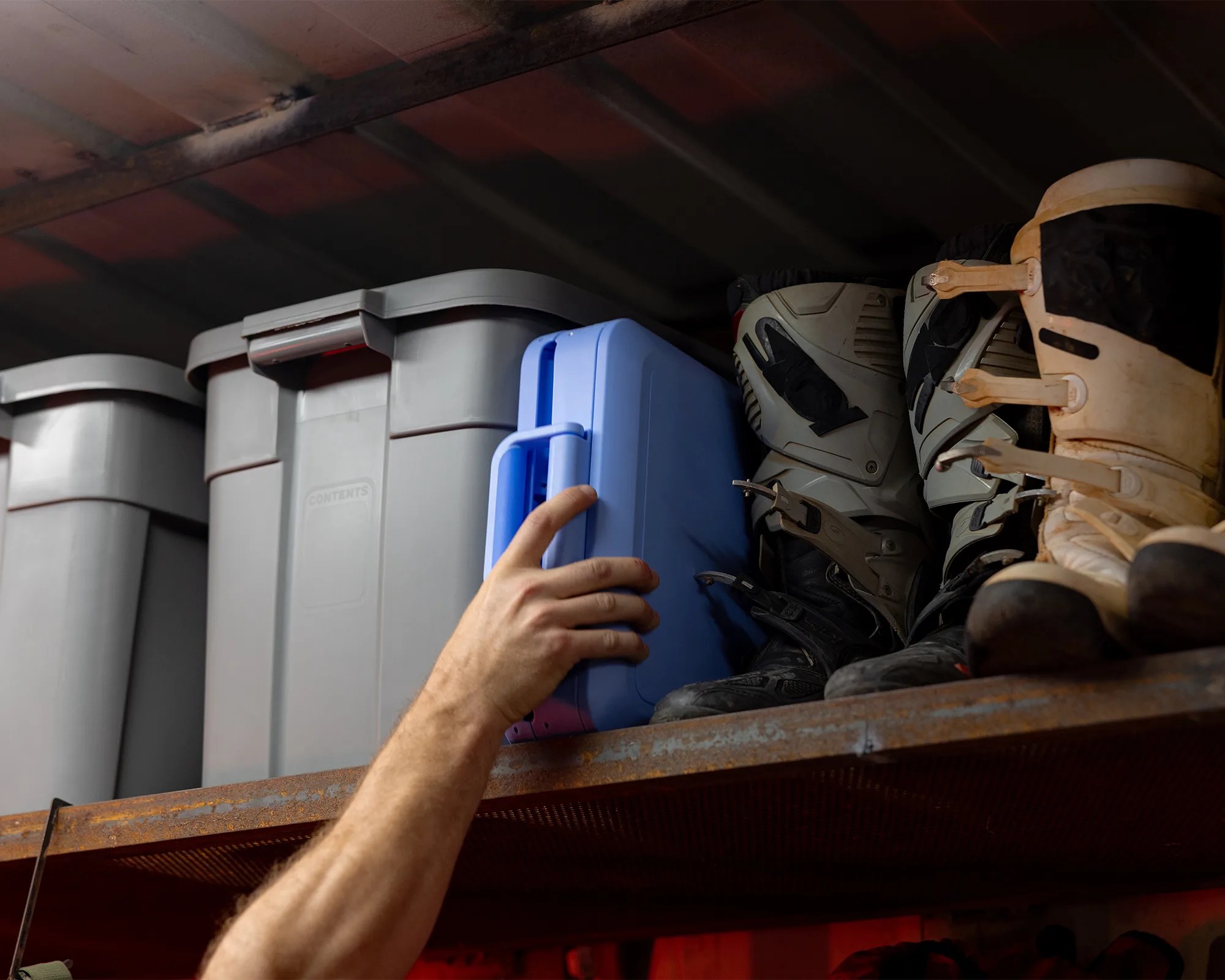 Hand reaching for a blue plastic storage container on a metal shelf next to gray bins and motocross boots.