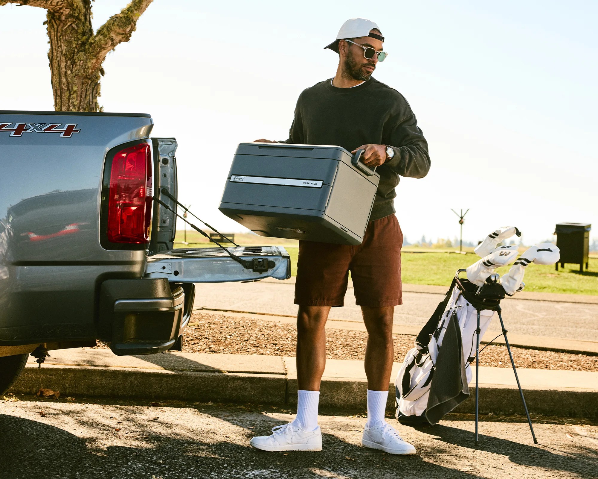 Man in sunglasses, white cap, black sweatshirt, and brown shorts loading a gray Coleman Snap 'N Go cooler into a pickup truck bed near a golf bag.