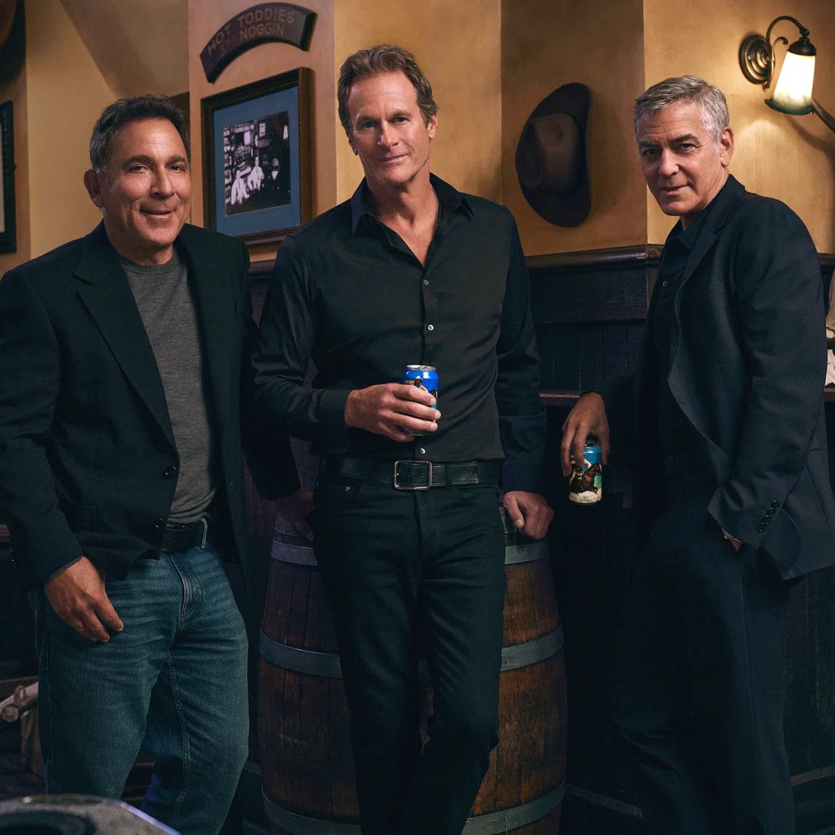 Three men in dark jackets and shirts holding cans, standing indoors near a wooden barrel and wall with framed pictures.