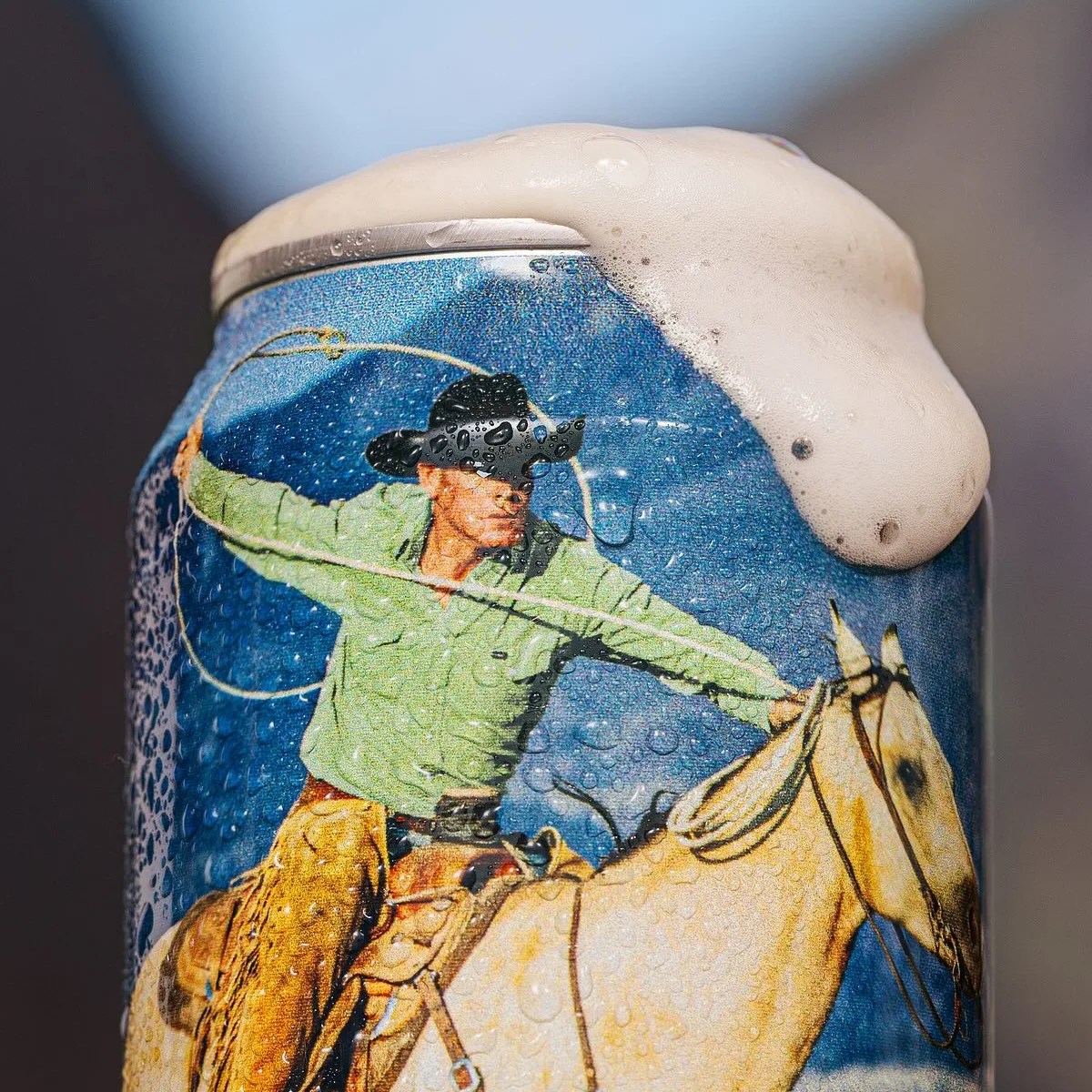Close-up of a wet beer can with foam overflowing, featuring a cowboy in a green shirt and black hat riding a horse.