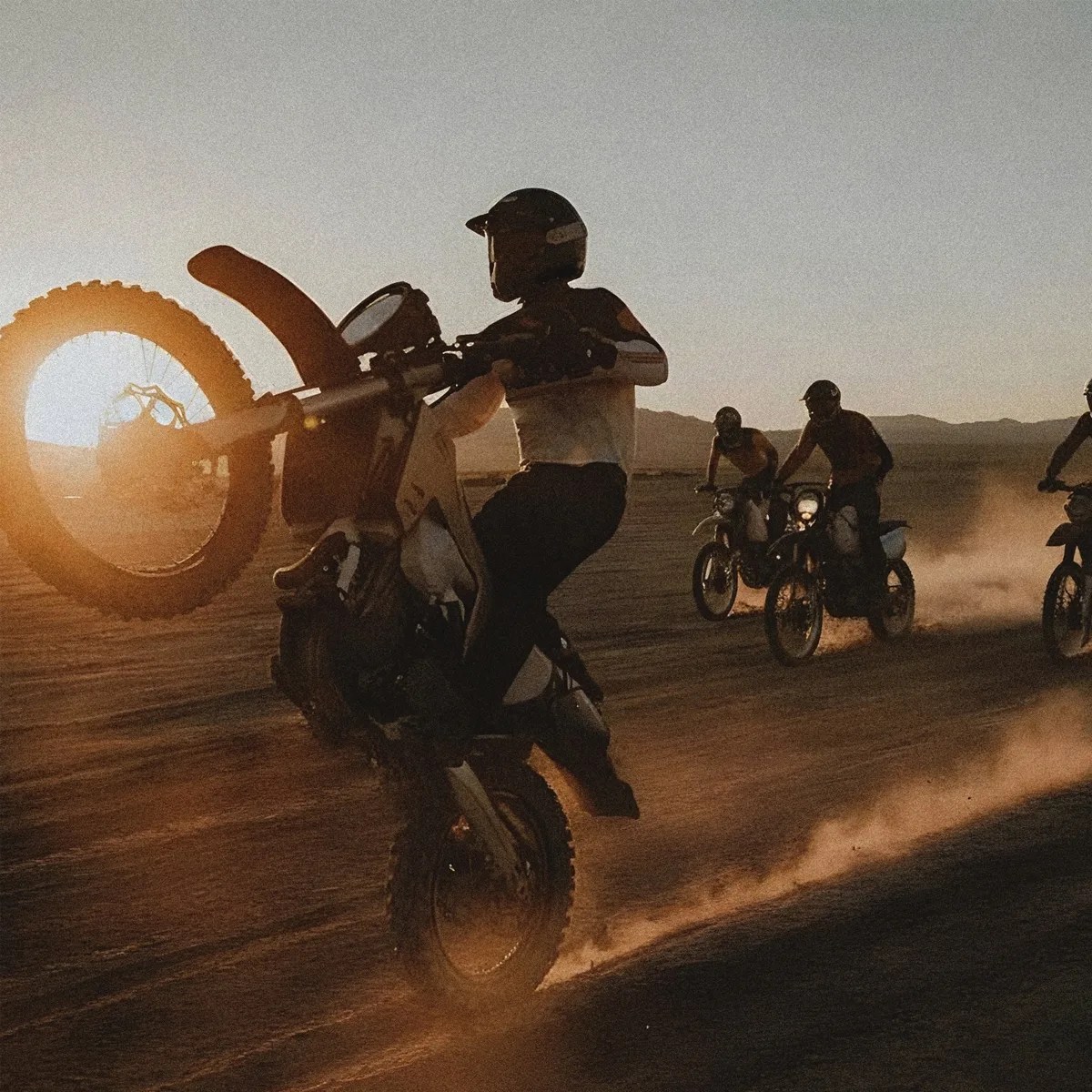 Motorcyclist performing a wheelie on a dirt bike at sunset with three other riders in the background on a dusty terrain.