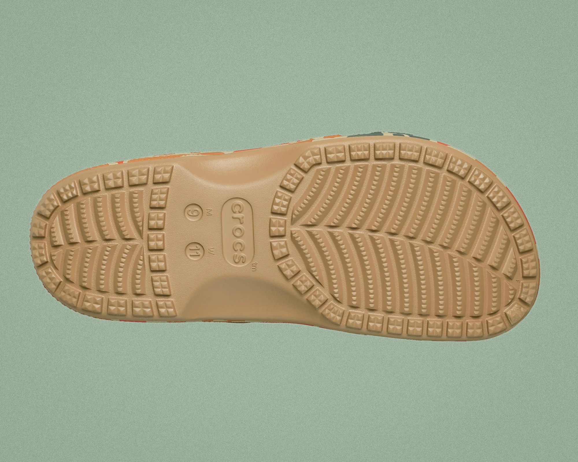 Light brown rubber sole of a Crocs shoe with textured tread and size markings on a green background.