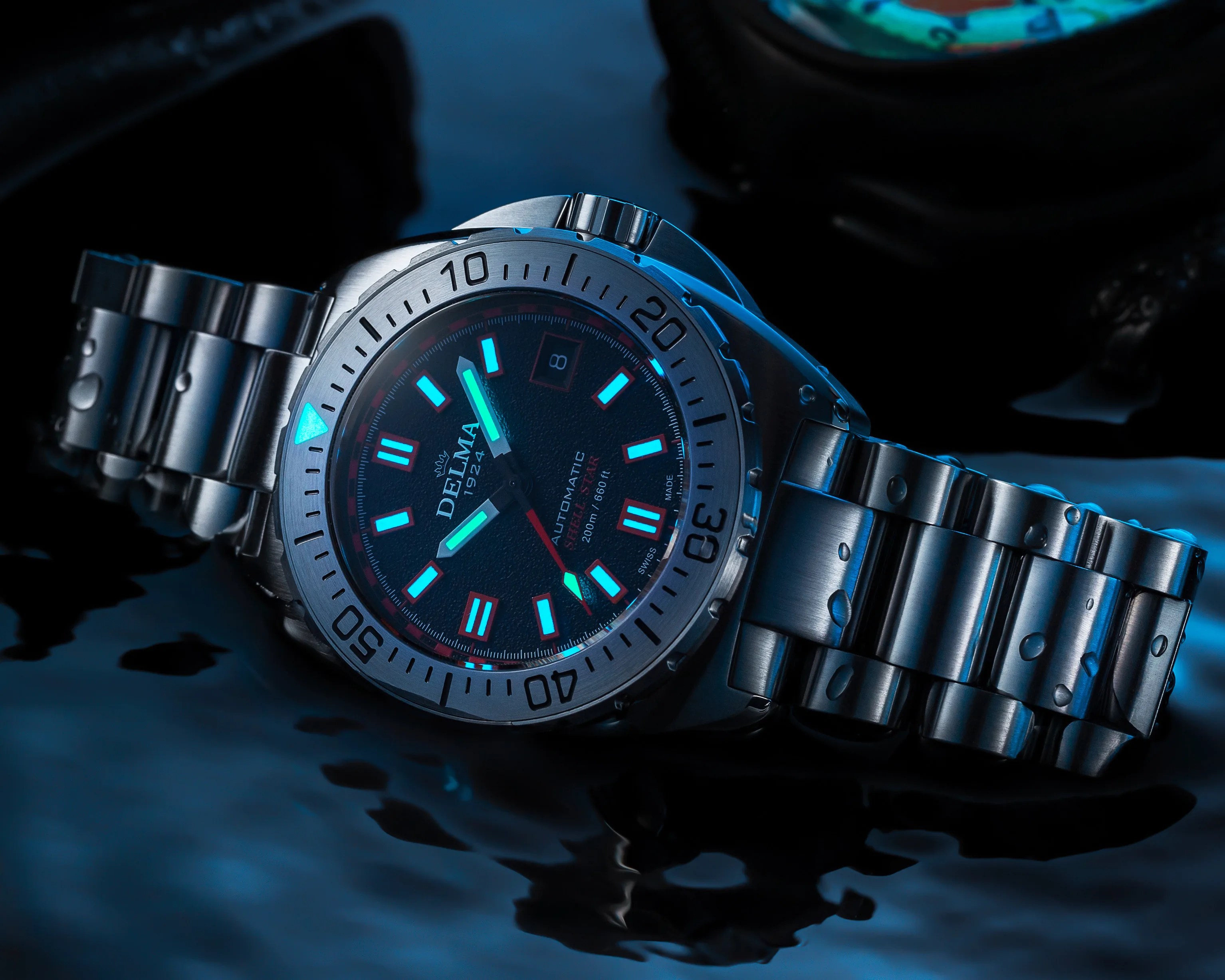 Stainless steel Delma automatic dive watch with luminous blue markers and water droplets on the bracelet.