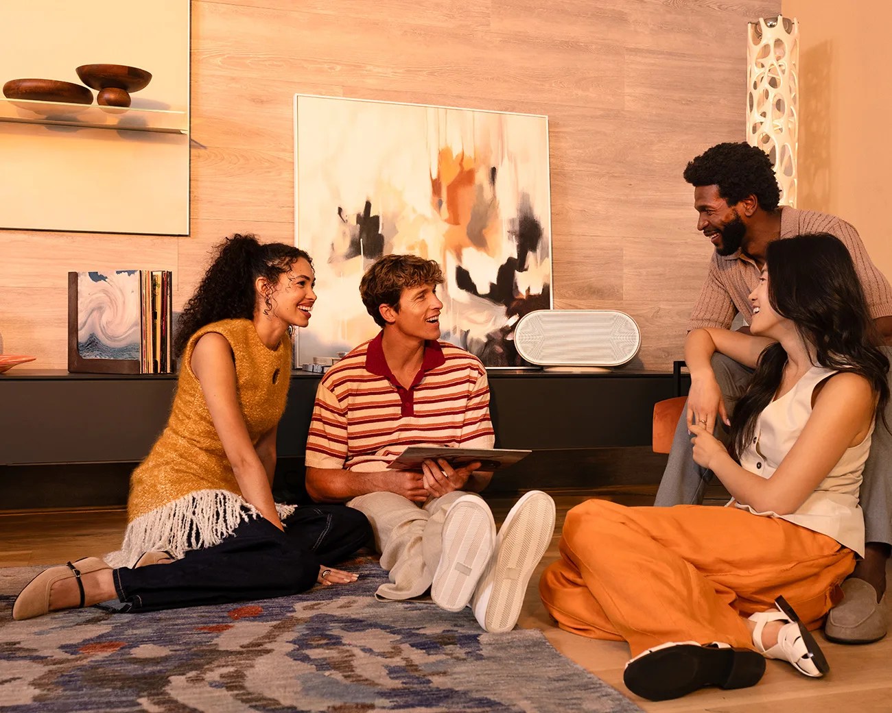 Four people sitting on a patterned rug in a modern living room with abstract art and a white speaker on a black cabinet.
