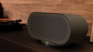 Dark gray fabric-covered Denon wireless speaker with rounded edges on a black surface.