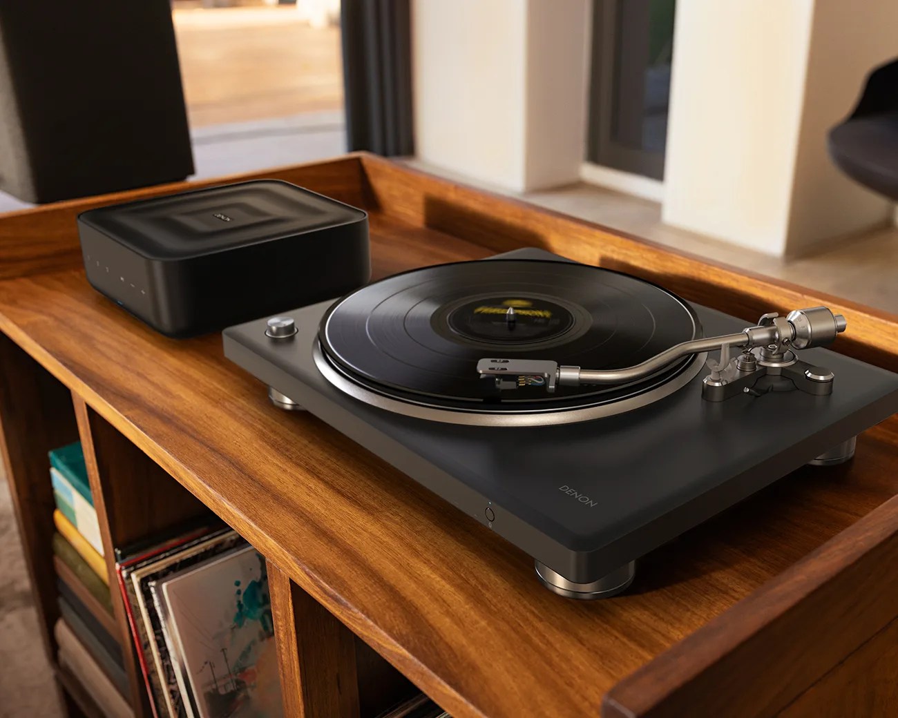 Black Denon turntable with silver tonearm playing a vinyl record on a wooden shelf with a black electronic device beside it.