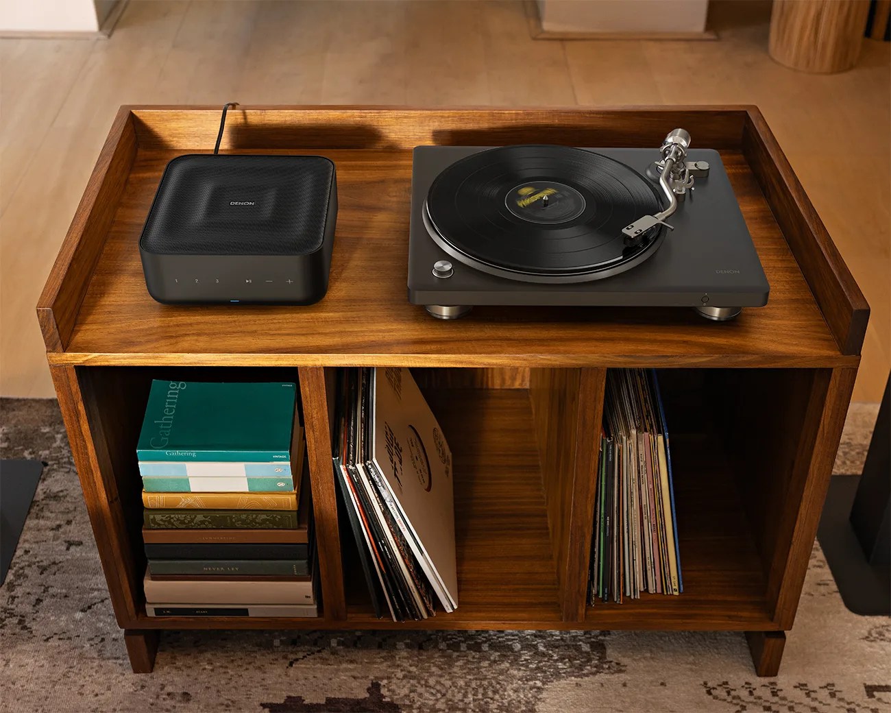 Wooden cabinet with three compartments holding books and vinyl records, topped with a black Denon turntable and speaker.