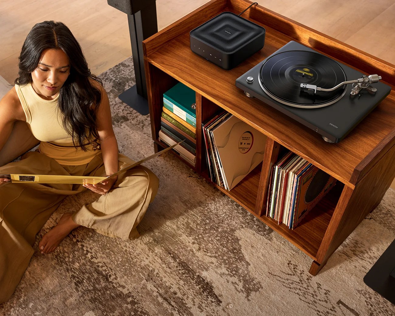 Woman sitting on a patterned rug holding a vinyl record near a wooden cabinet with a Denon turntable and speaker.