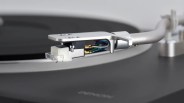 Close-up of a silver tonearm and cartridge with colored wires on a black turntable.