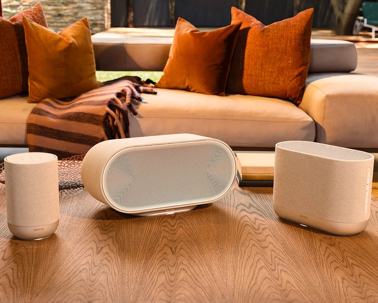 Three beige Denon wireless speakers of different shapes on a wooden table in front of a beige sofa with orange pillows and a striped blanket.