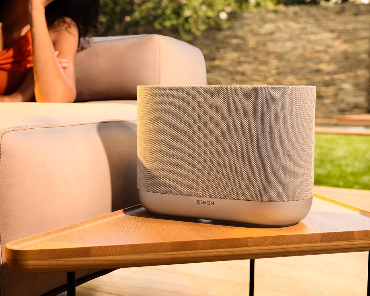 Beige Denon wireless speaker with fabric cover on a wooden table next to a beige sofa.