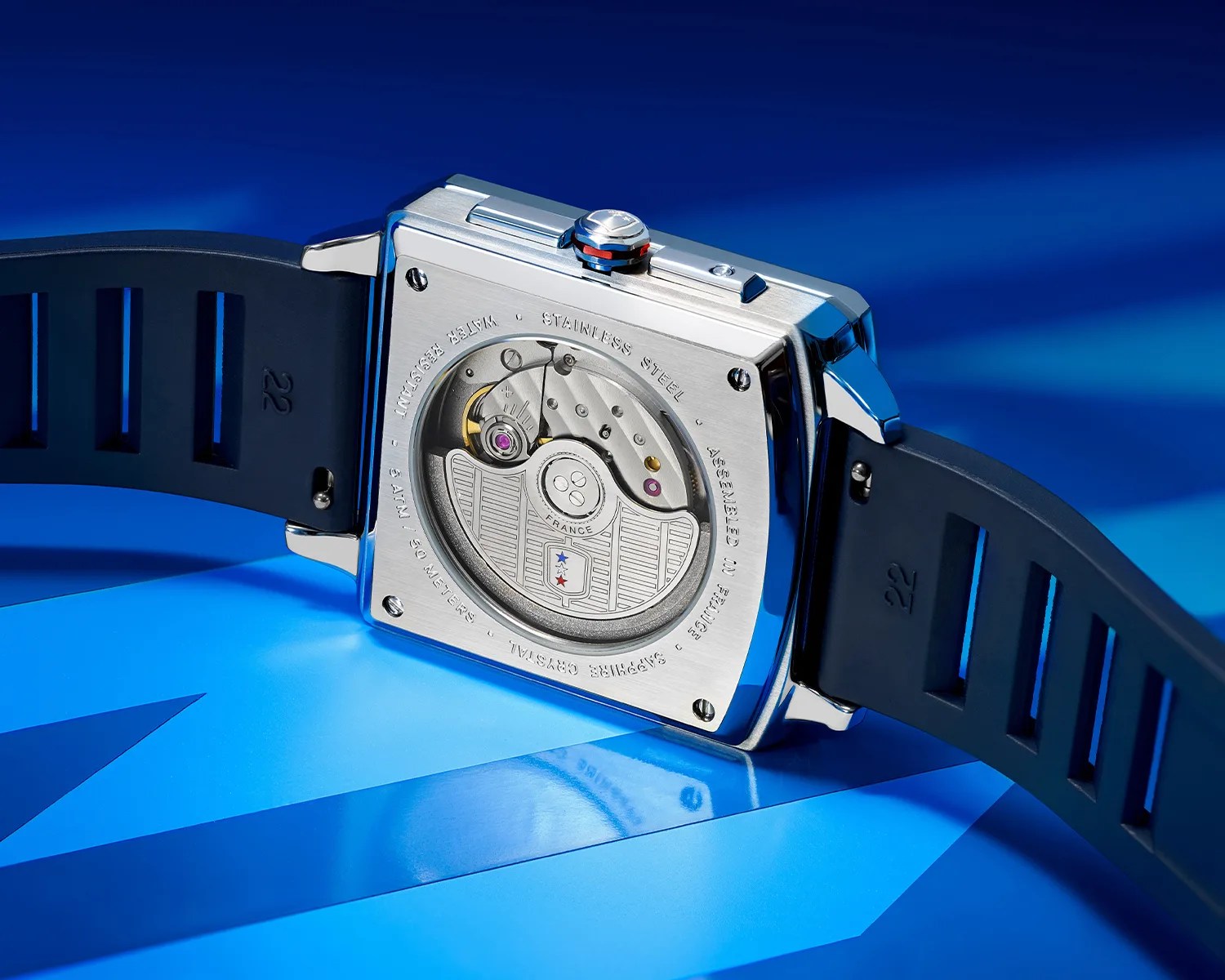 Square stainless steel watch case back with visible automatic movement and black rubber strap marked "22".