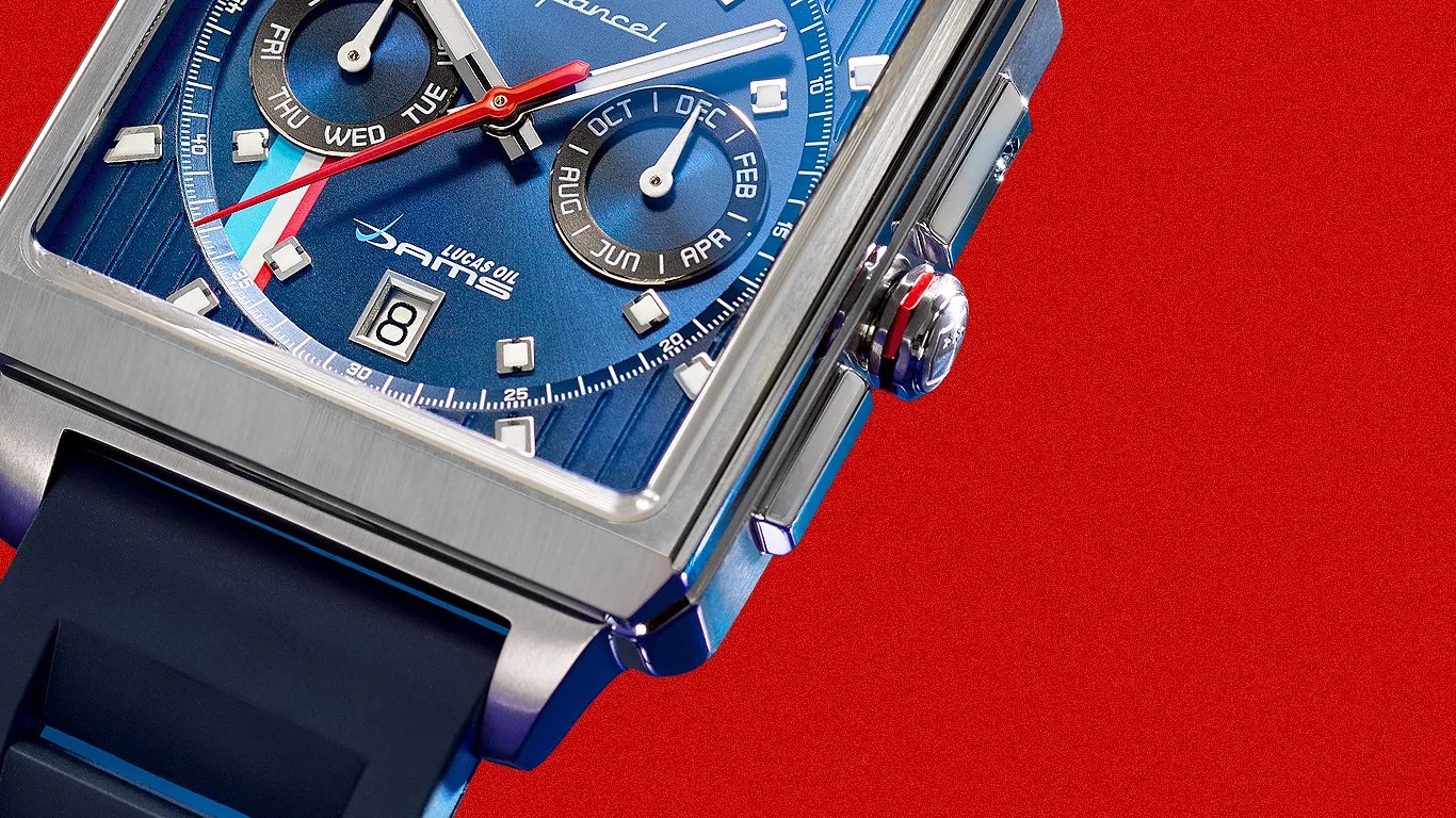 Square stainless steel watch with blue dial, subdials, date window, and black rubber strap on red background.