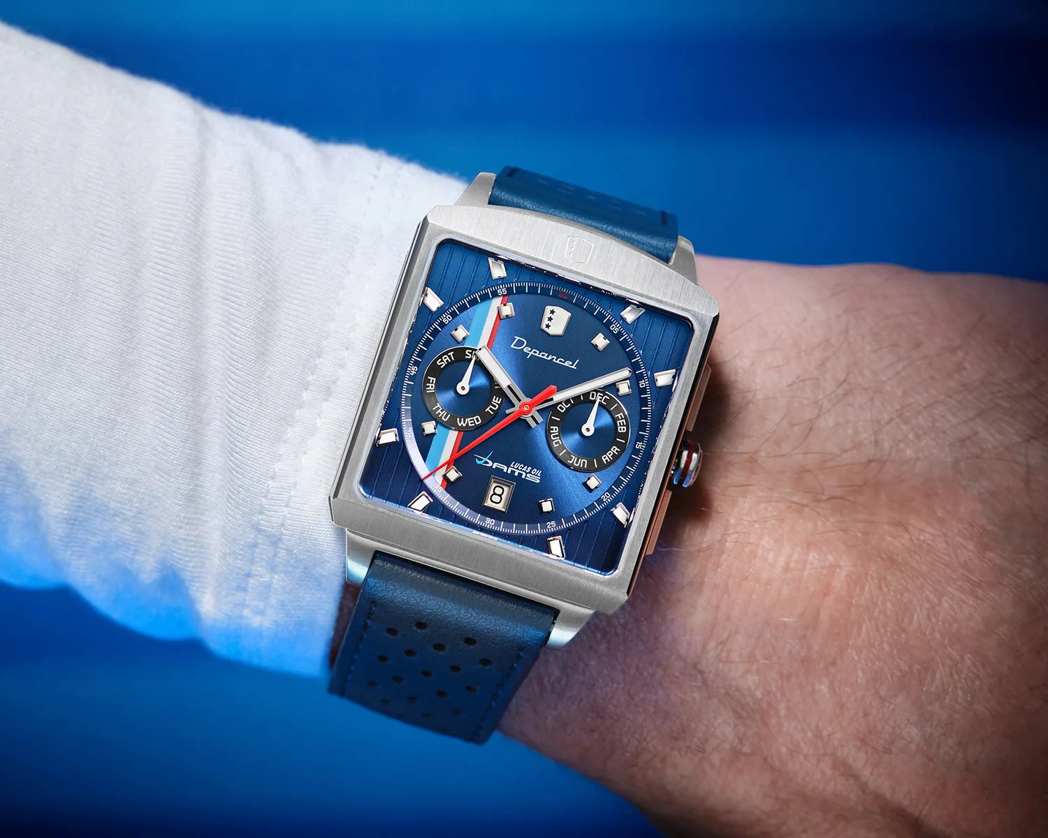 Square silver wristwatch with blue dial, red and white hands, and blue perforated leather strap worn on a wrist.