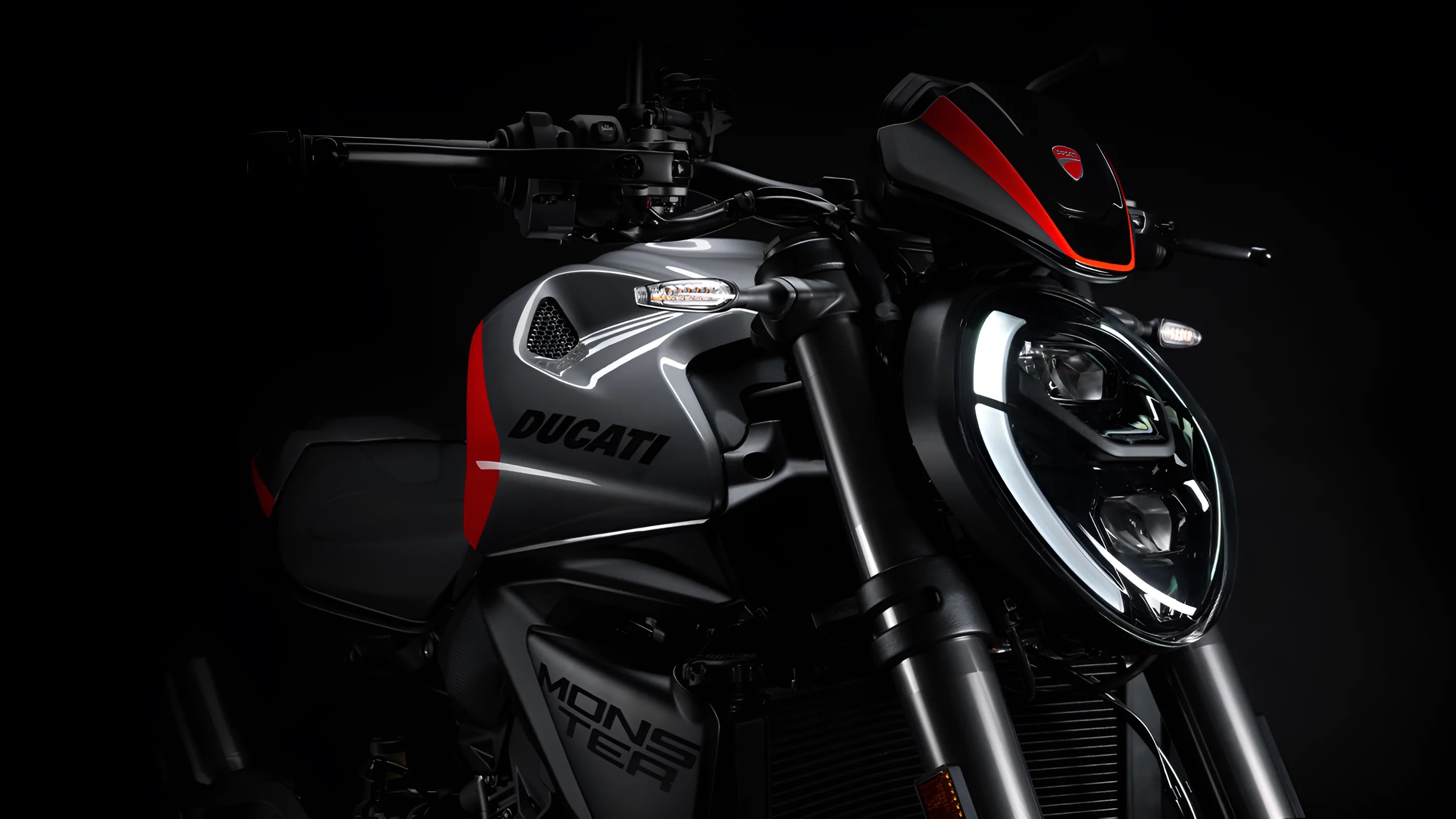 Close-up of a black and red Ducati Monster motorcycle front with illuminated LED headlight and visible brand logos.