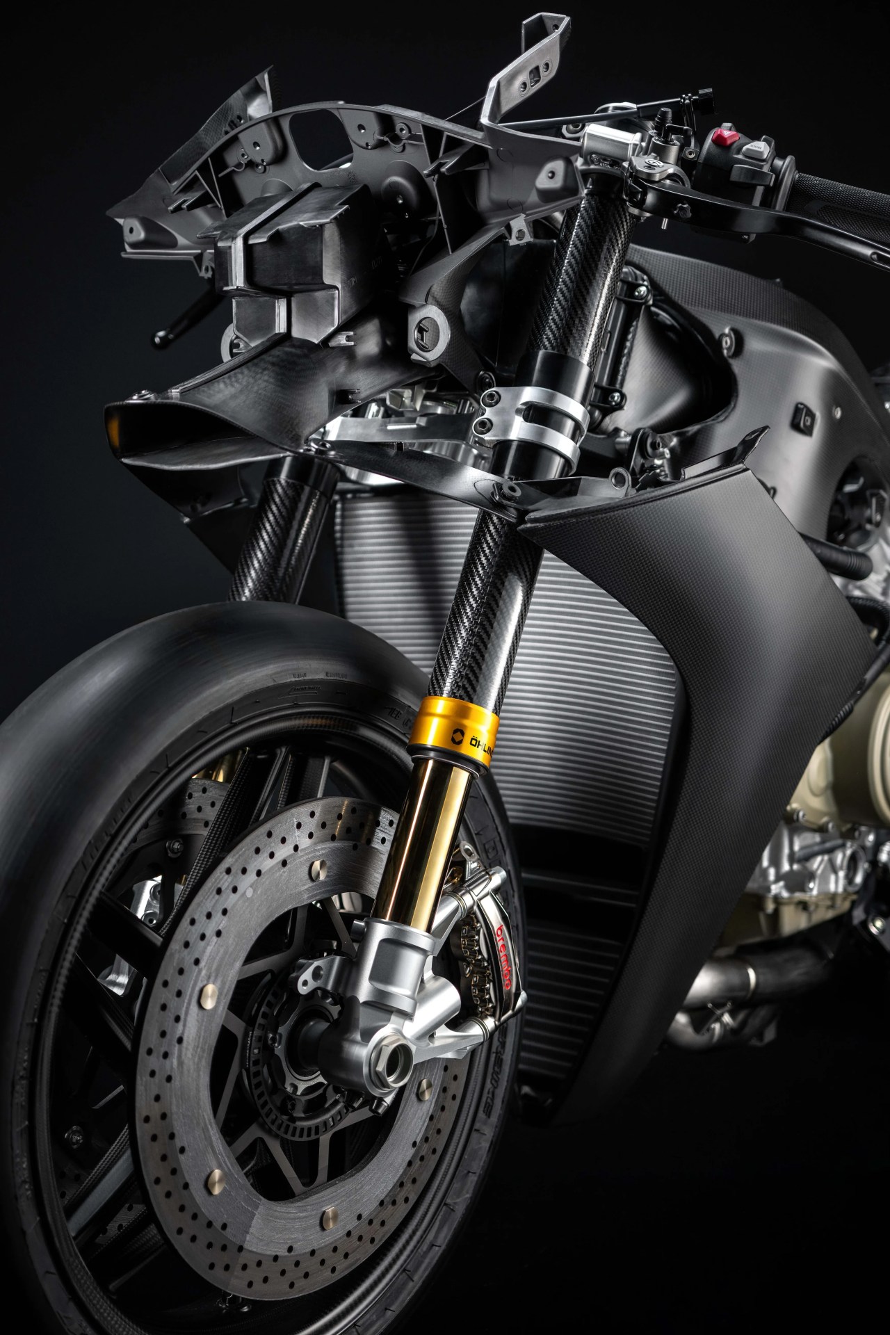 Close-up of a motorcycle front suspension with carbon fiber forks, gold accents, and a Brembo brake caliper.