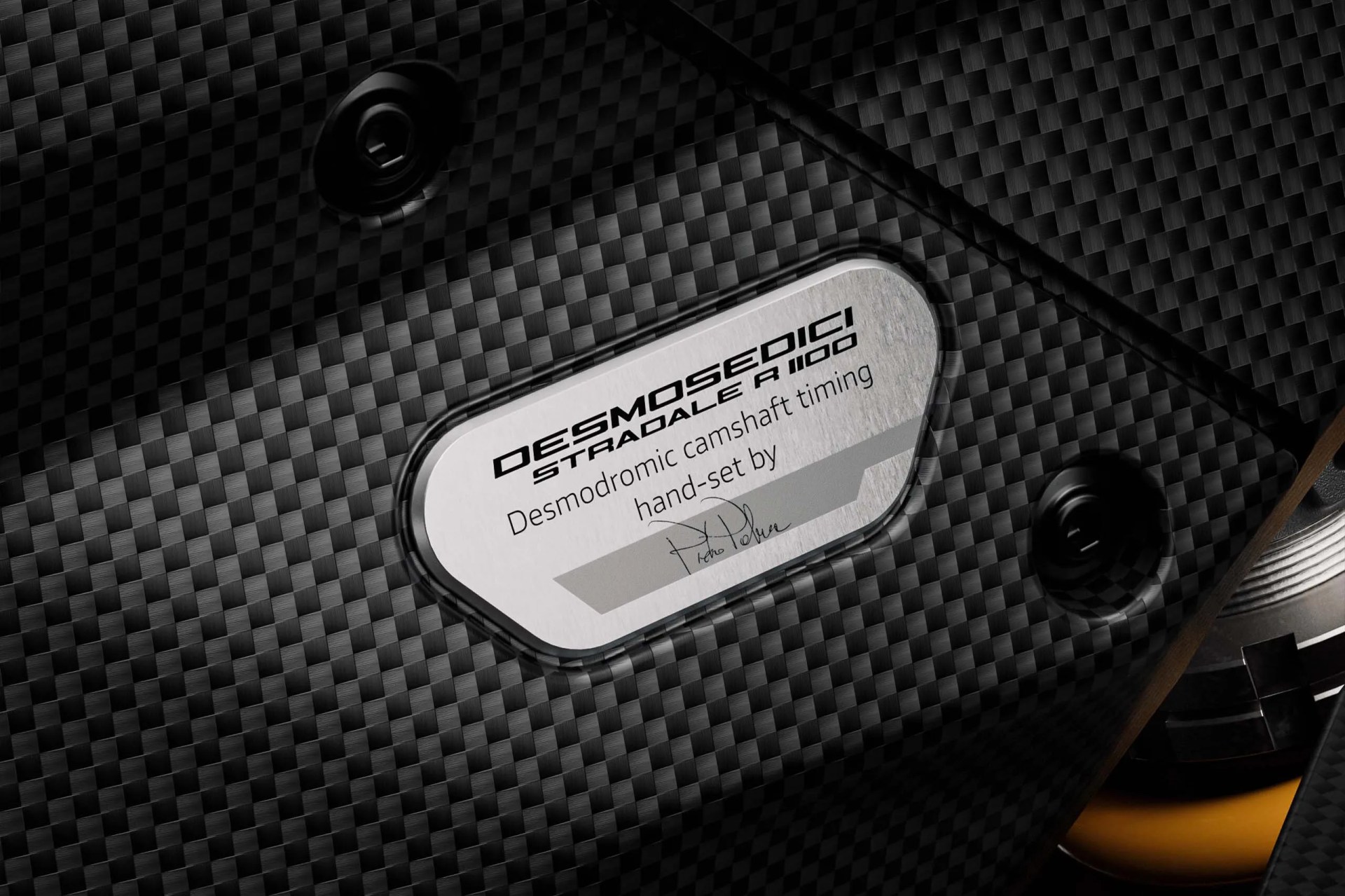 Close-up of a carbon fiber engine cover with a metal plaque reading "Desmosedici Stradale R 1100 Desmodromic camshaft timing hand-set by" and a signature.