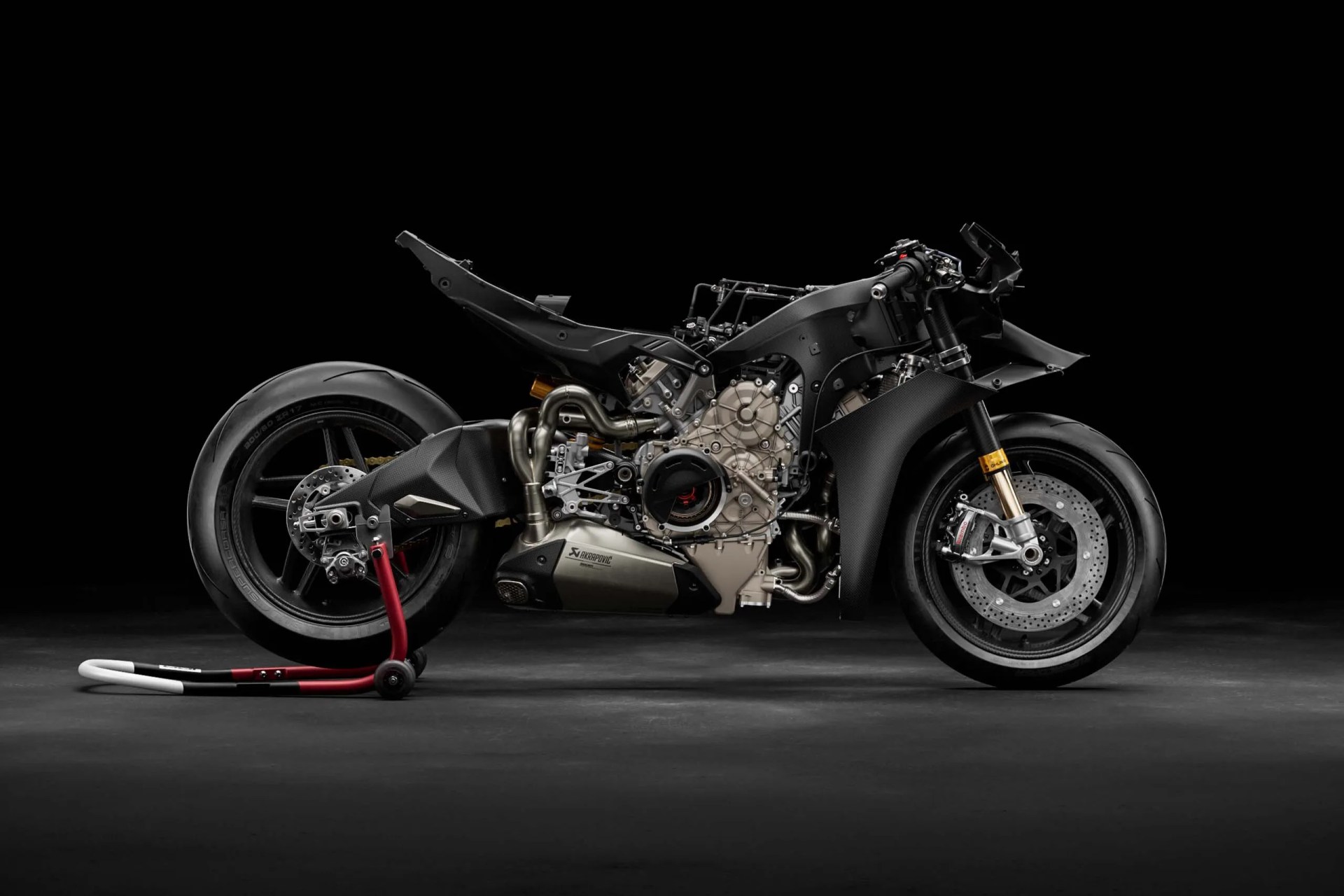 Black racing motorcycle frame with exposed engine and exhaust on a rear stand against a black background.