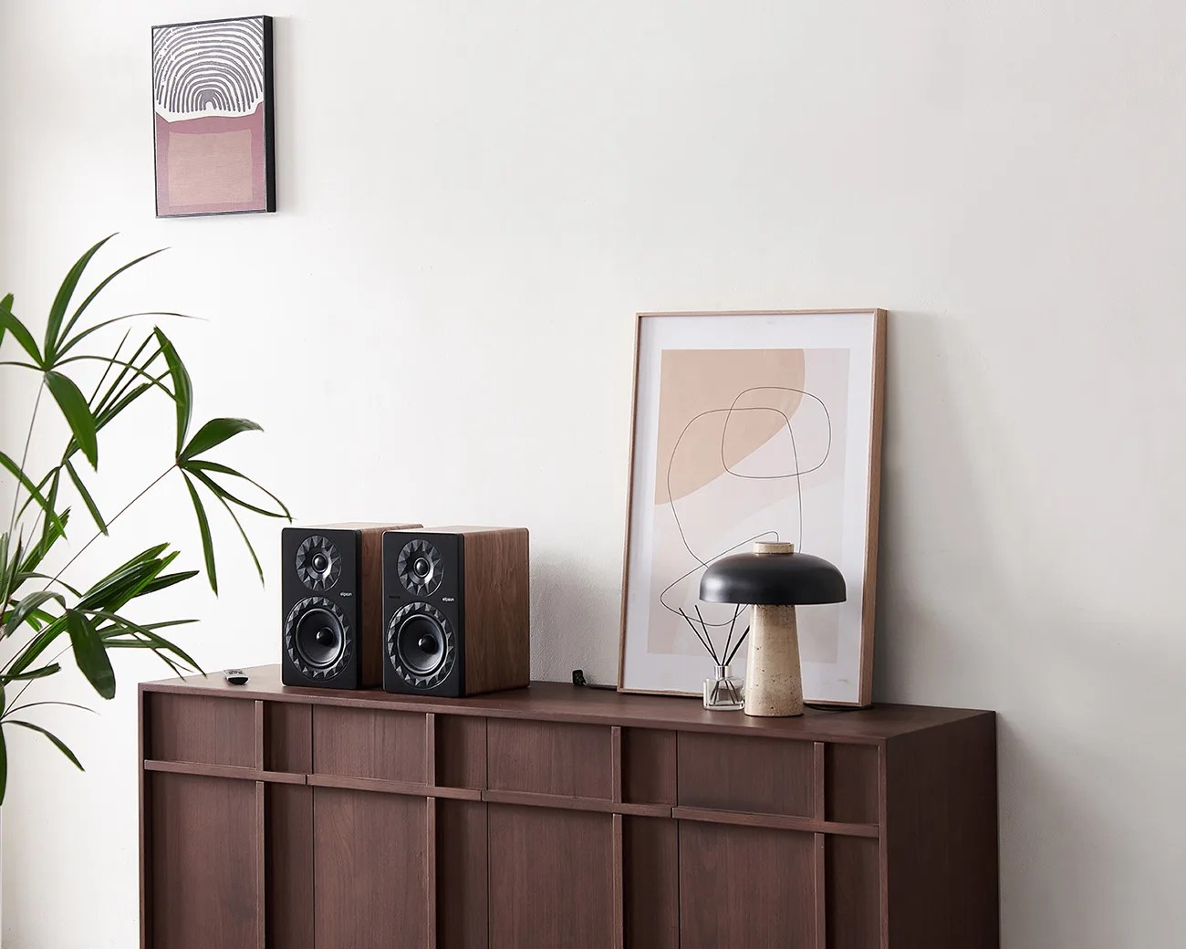Pair of black and wood bookshelf speakers on a dark wood cabinet with abstract art and a black mushroom-shaped lamp.