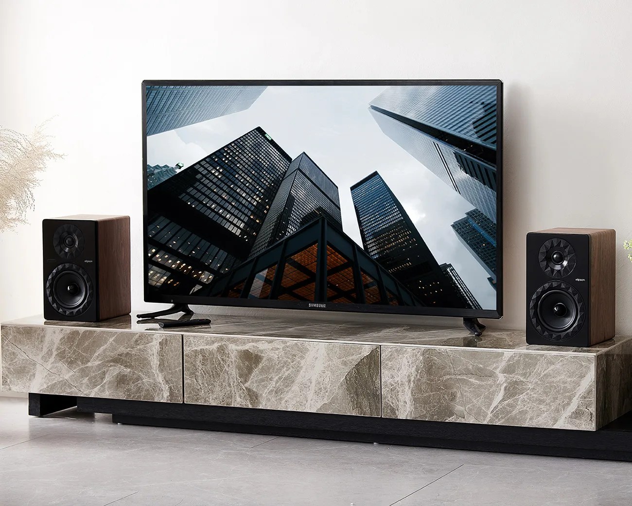 Samsung flat-screen TV displaying skyscrapers on a marble TV stand with two wooden bookshelf speakers.