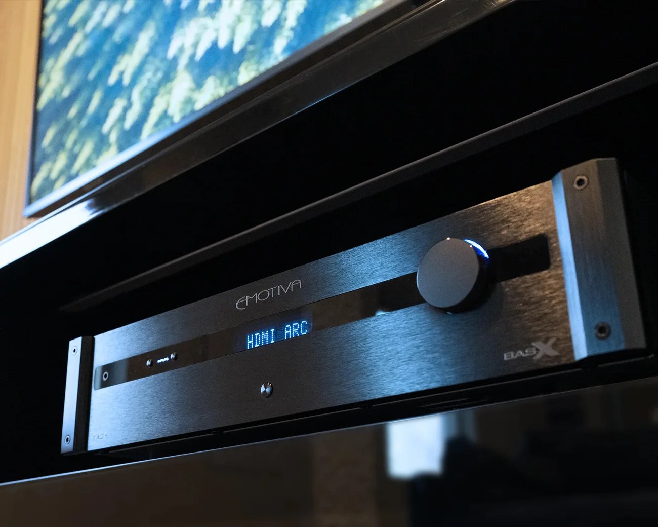 Black Emotiva audio receiver with a blue digital display showing "HDMI ARC" and a large volume knob.