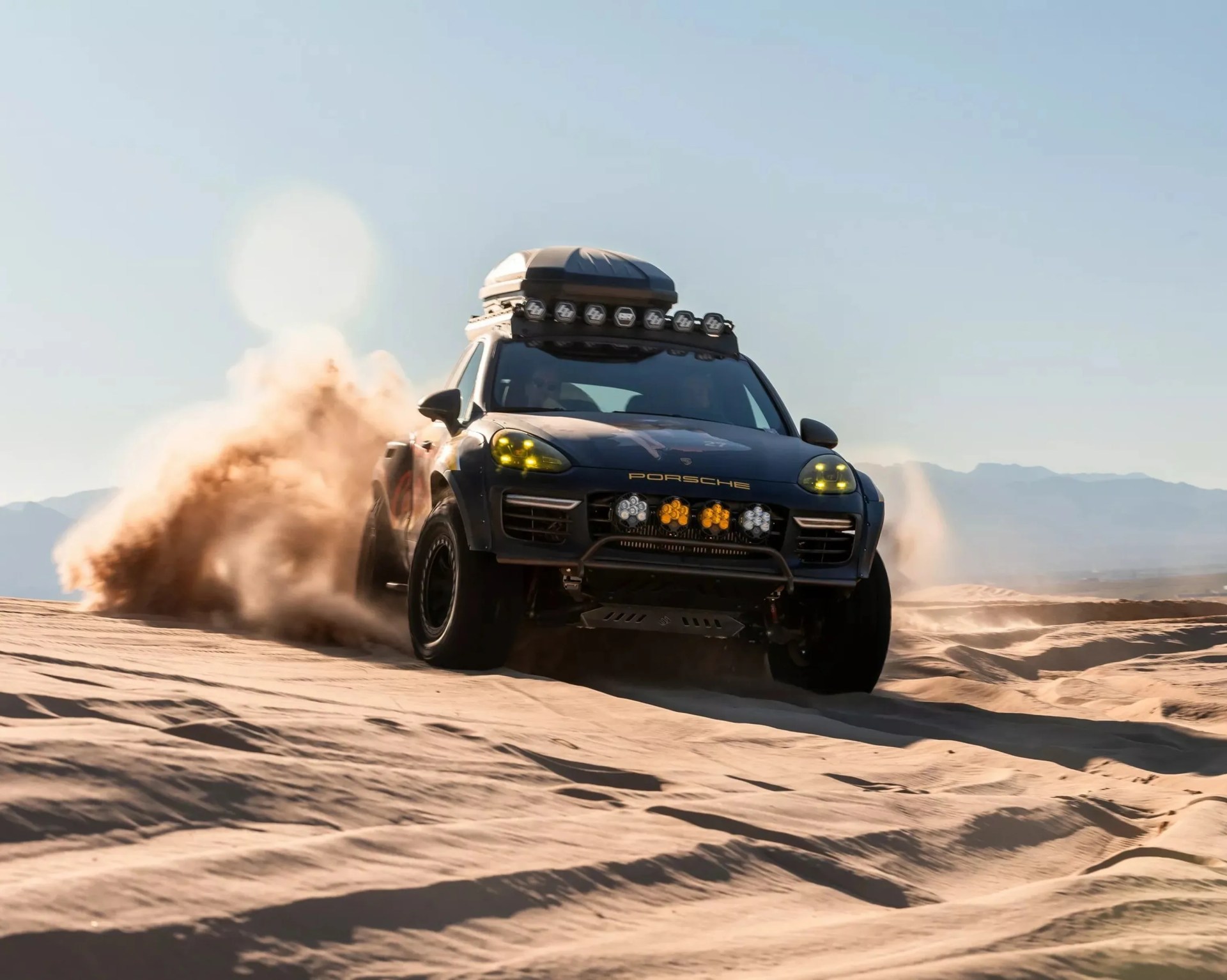 Black Porsche SUV with roof cargo box and off-road lights kicking up sand in a desert setting.